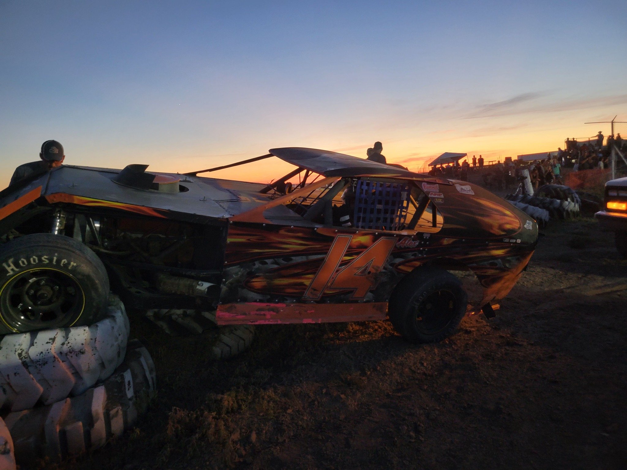 A dirt racetrack at sunset with race cars and spectators in the background. The focus is on a black racing car with orange and yellow flame designs, large tires, and a roll cage.