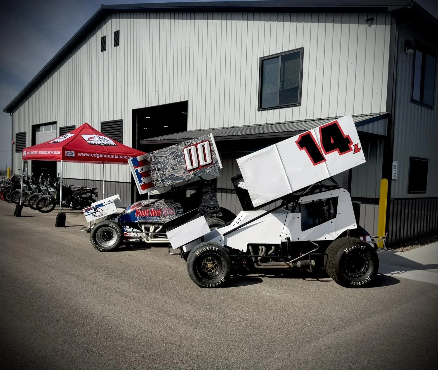 Two race cars with large rear wings parked outside a building, with a red canopy and motorcycles in the background.