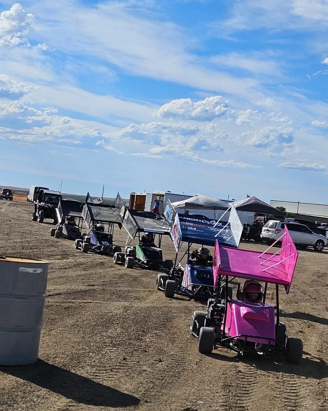 Line of small race cars with colorful canopies parked on dirt under a partly cloudy sky.