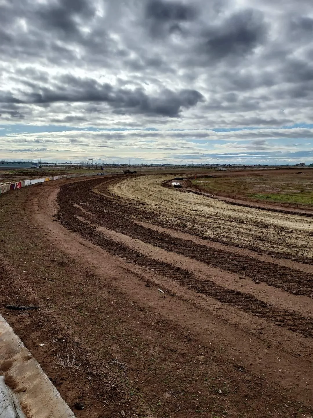 An empty dirt racetrack with tire marks and a muddy section, under a cloudy sky with patches of blue.