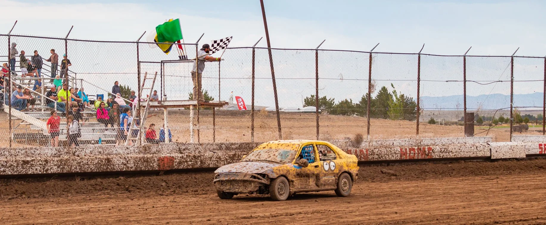A dirt track racing field with a yellow race car covered in dirt and mud. Spectators sit on bleachers behind a chain-link fence, while a person stands on a platform holding a checkered flag and a green flag, signaling the end of the race. The backgro