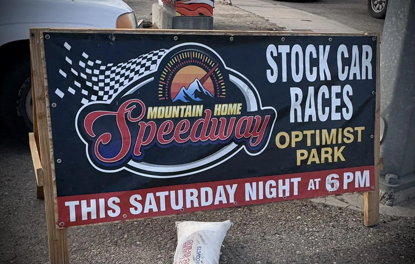 Signboard advertising Mountain Speedway with details about stock car races and a race event on Saturday at 6 PM. The sign features a logo with mountains and a sunset, checkered flag graphic, and bold text.