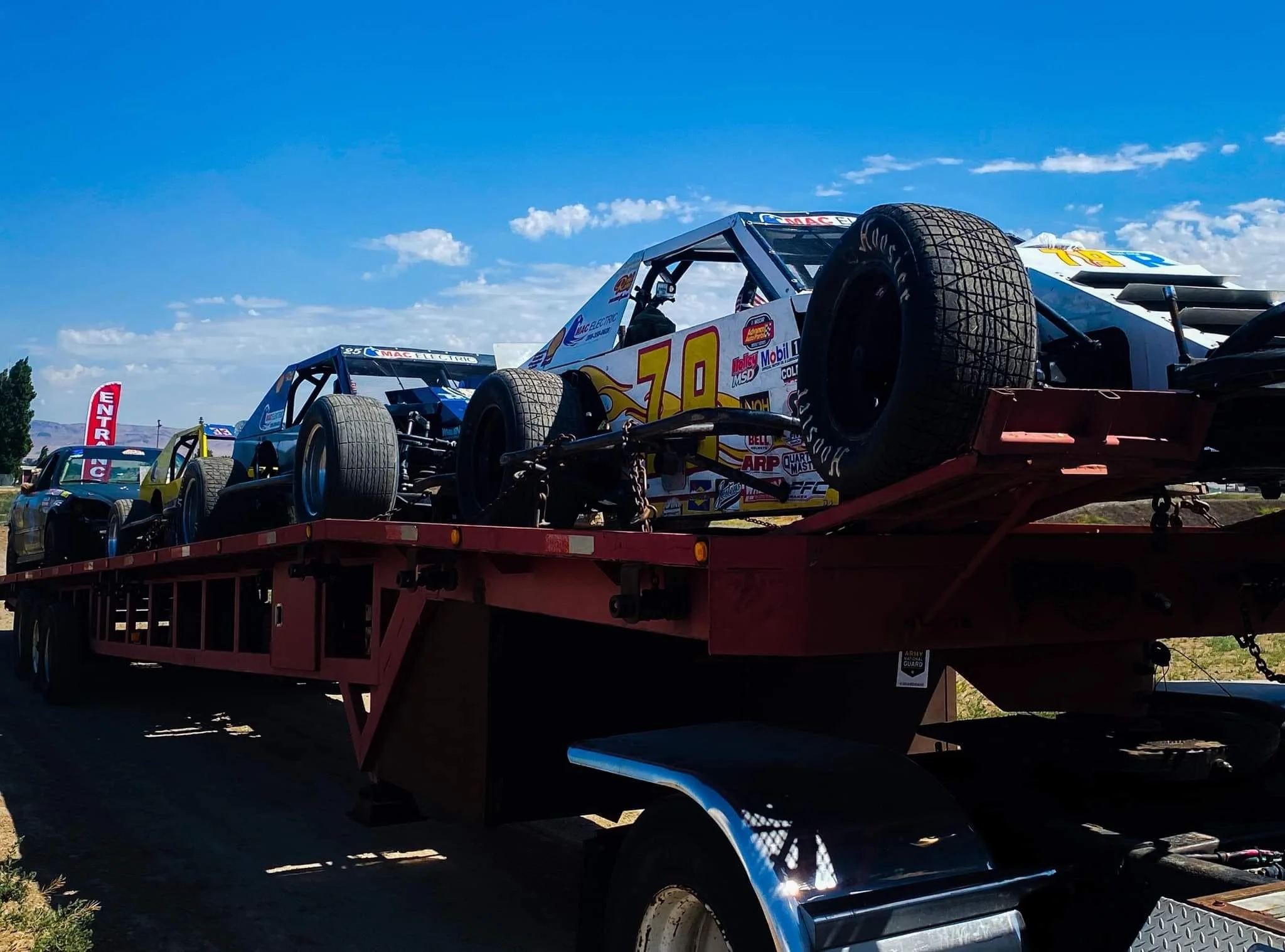 Race cars loaded on a red flatbed trailer under a blue sky with scattered clouds.