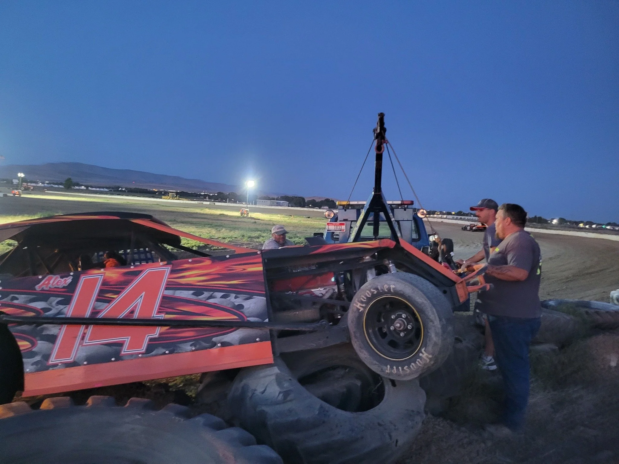 A race car with the number 4 on it, parked on a dirt track at dusk, with a group of people gathered around, some wearing caps and safety vests, and race trailers and lights visible in the background.