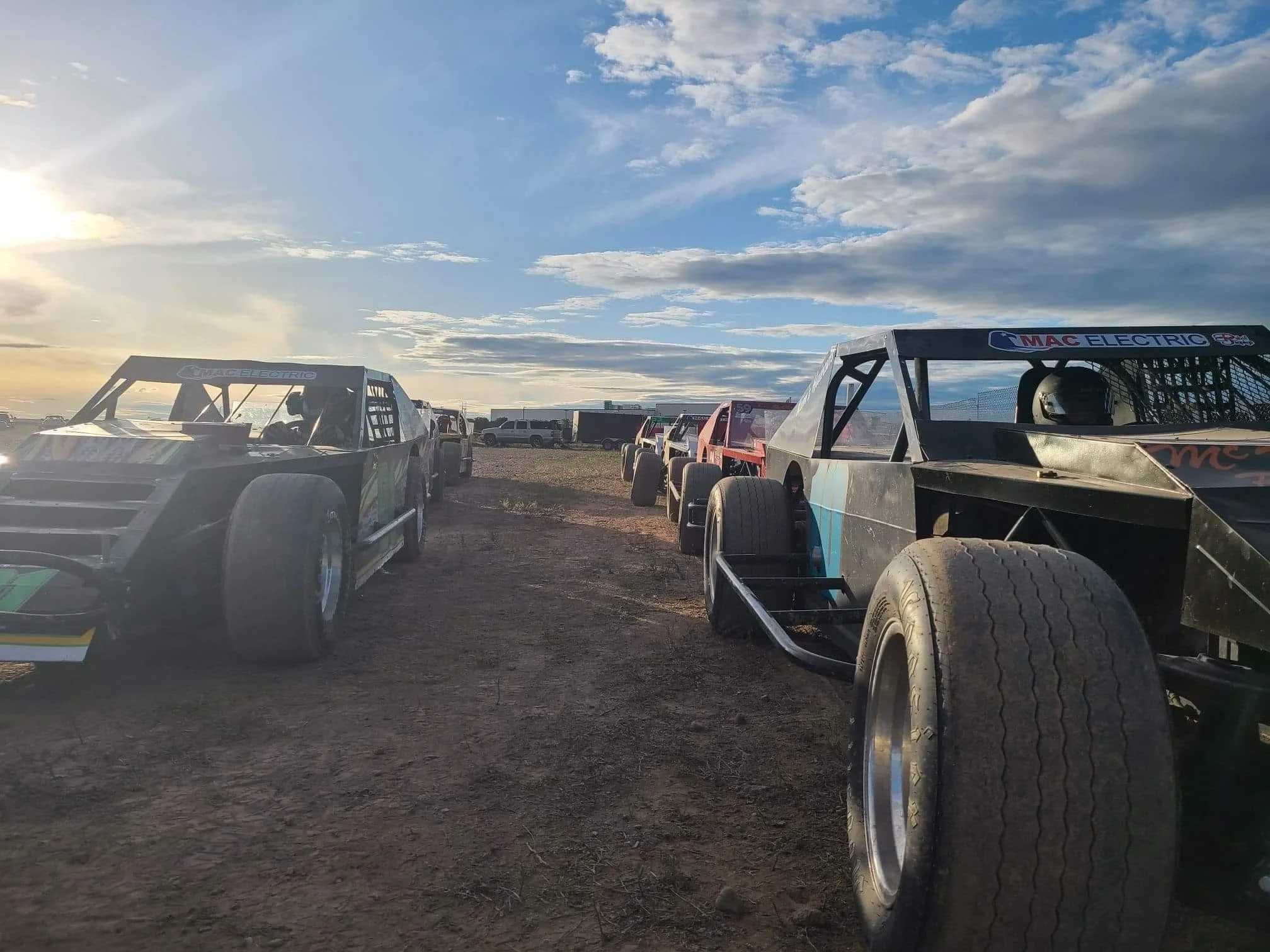 Race cars parked on a dirt lot during sunset, with a partly cloudy sky above.