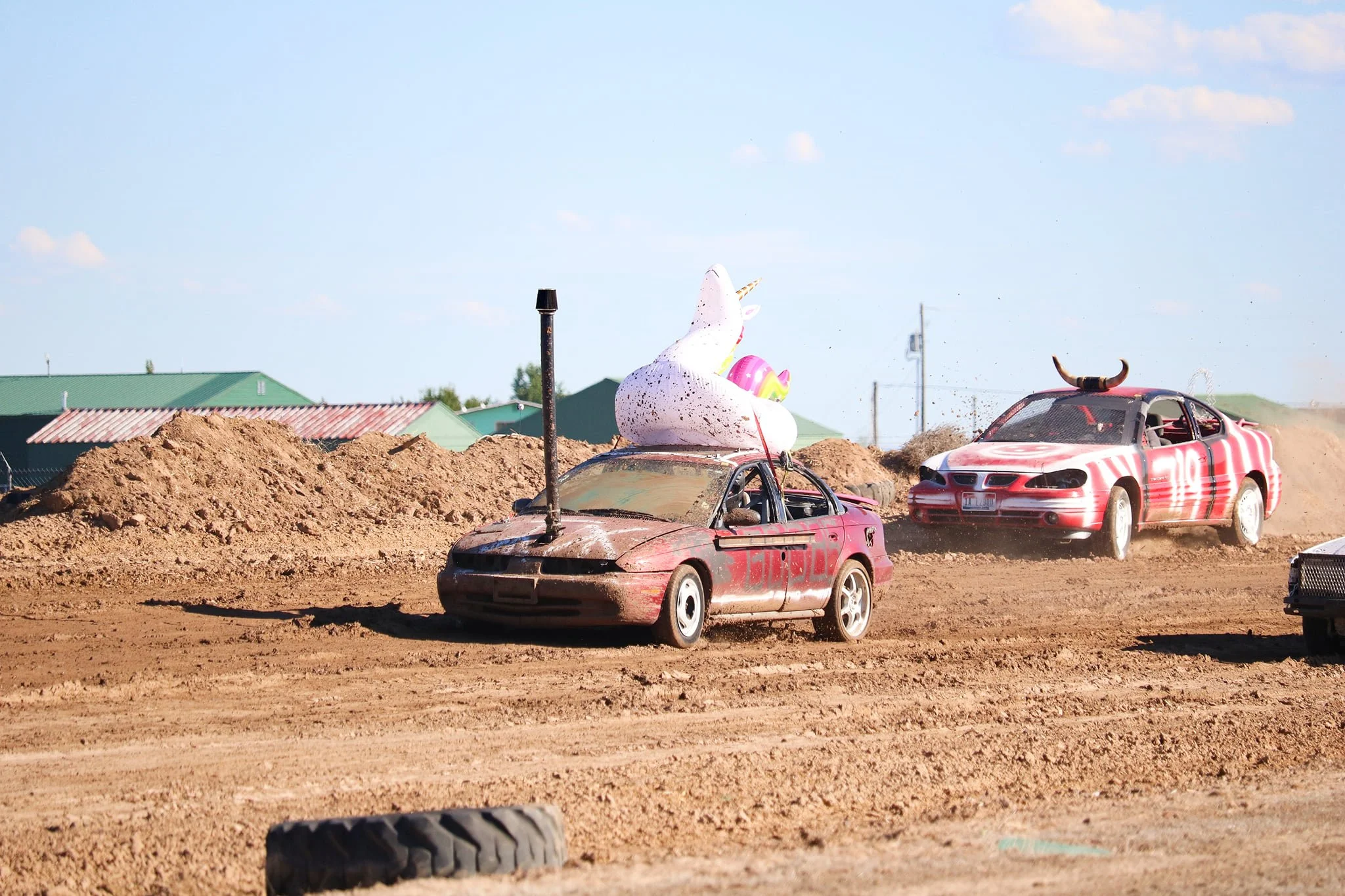 Several old, rusty cars with carnival decorations on top are racing on a dirt track during daytime. The cars are covered in dirt and dust, and the background features a dirt mound, green-roofed buildings, and a few utility poles under a blue sky with