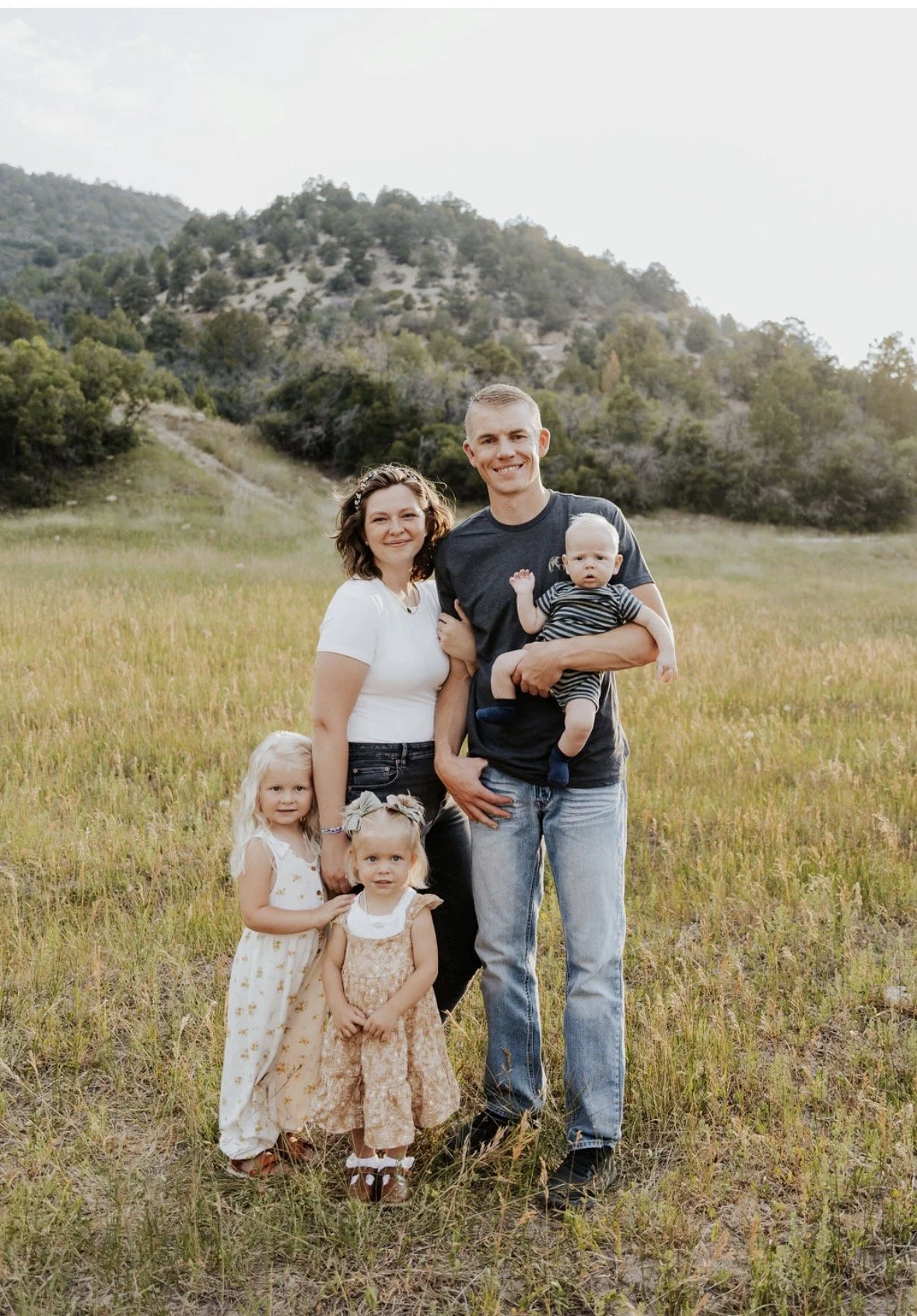 A family of six standing on a grassy field outdoors with mountains and trees in the background, smiling at the camera. The family includes two adults and four children, three girls and one boy.