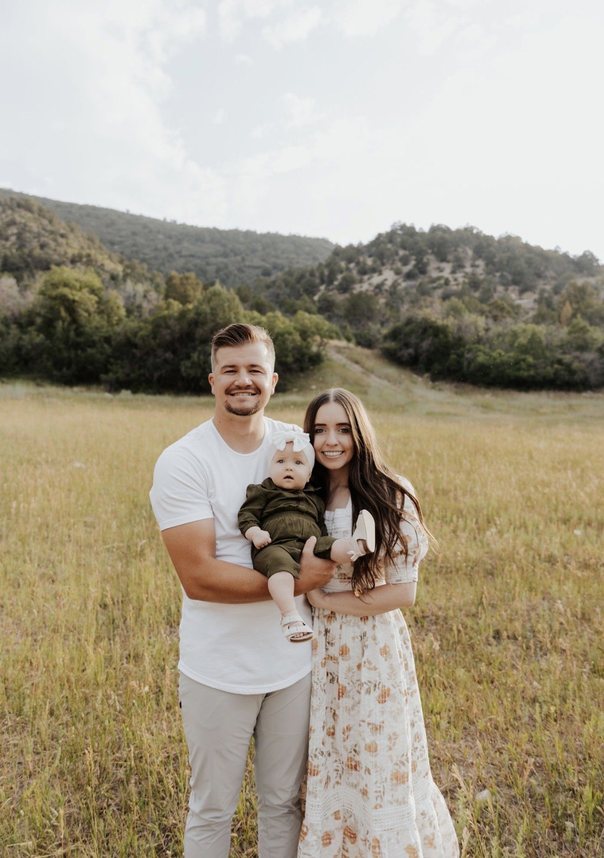 A family of three standing in a grassy field with mountains and trees in the background. The man is holding a baby girl, and the woman is standing beside them. All are smiling.
