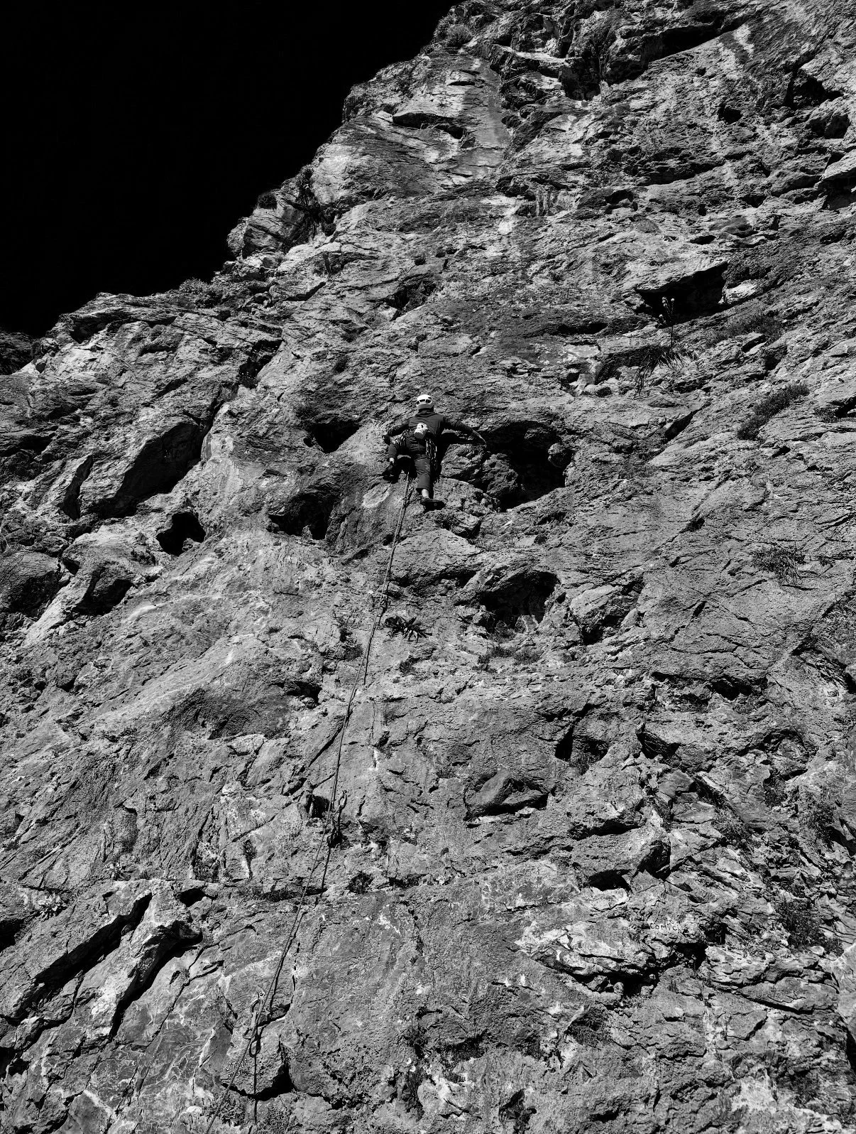 A person climbing a steep, rocky mountain face with safety gear, ropes, and a helmet in a black-and-white photograph.