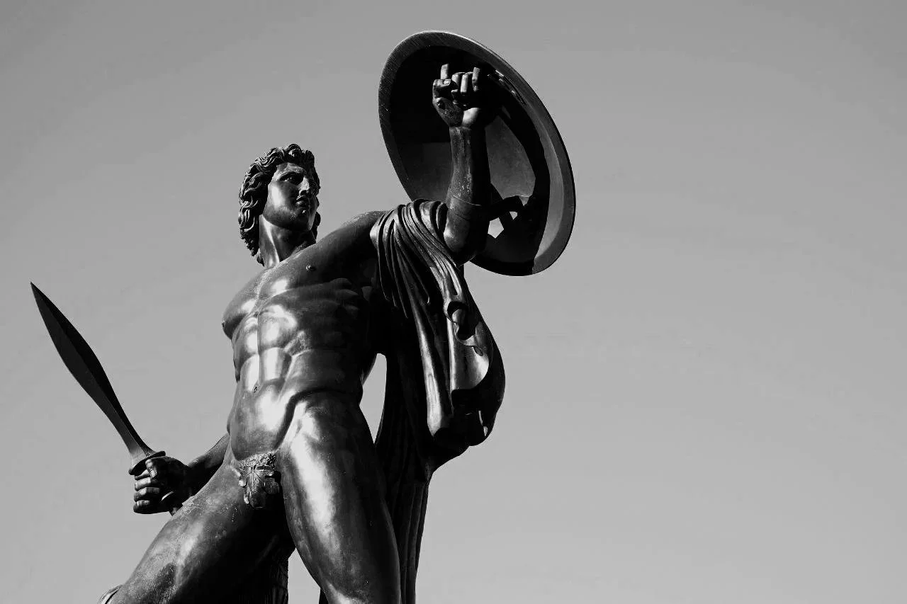 Black and white sculpture of a muscular man holding a sword in one hand and a shield in the other, set against a clear sky