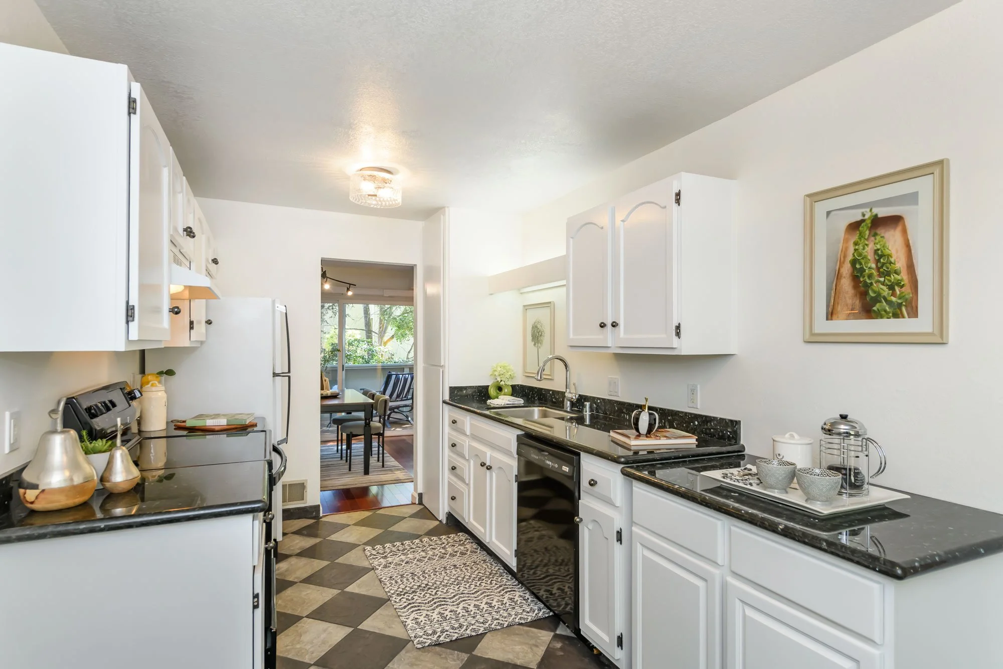 A kitchen with white cabinets and black countertops, with a view into a dining area and living room with large windows and trees outside.