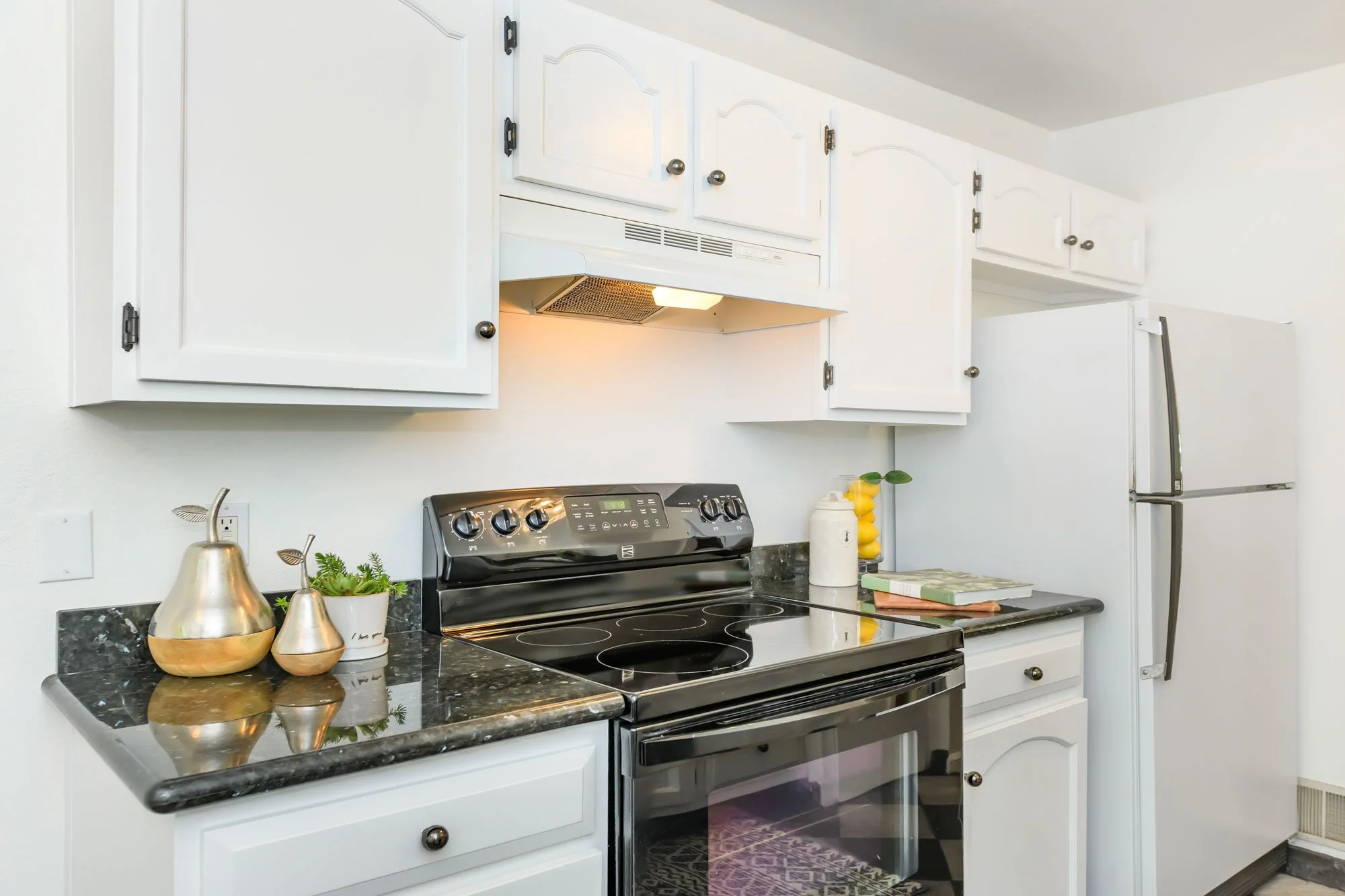 White kitchen cabinets above a black electric stove with a marbled black countertop. Decorative gold and white vases, a small potted plant, and a stack of books are on the countertop. To the right is a white appliance, possibly a refrigerator, and a 