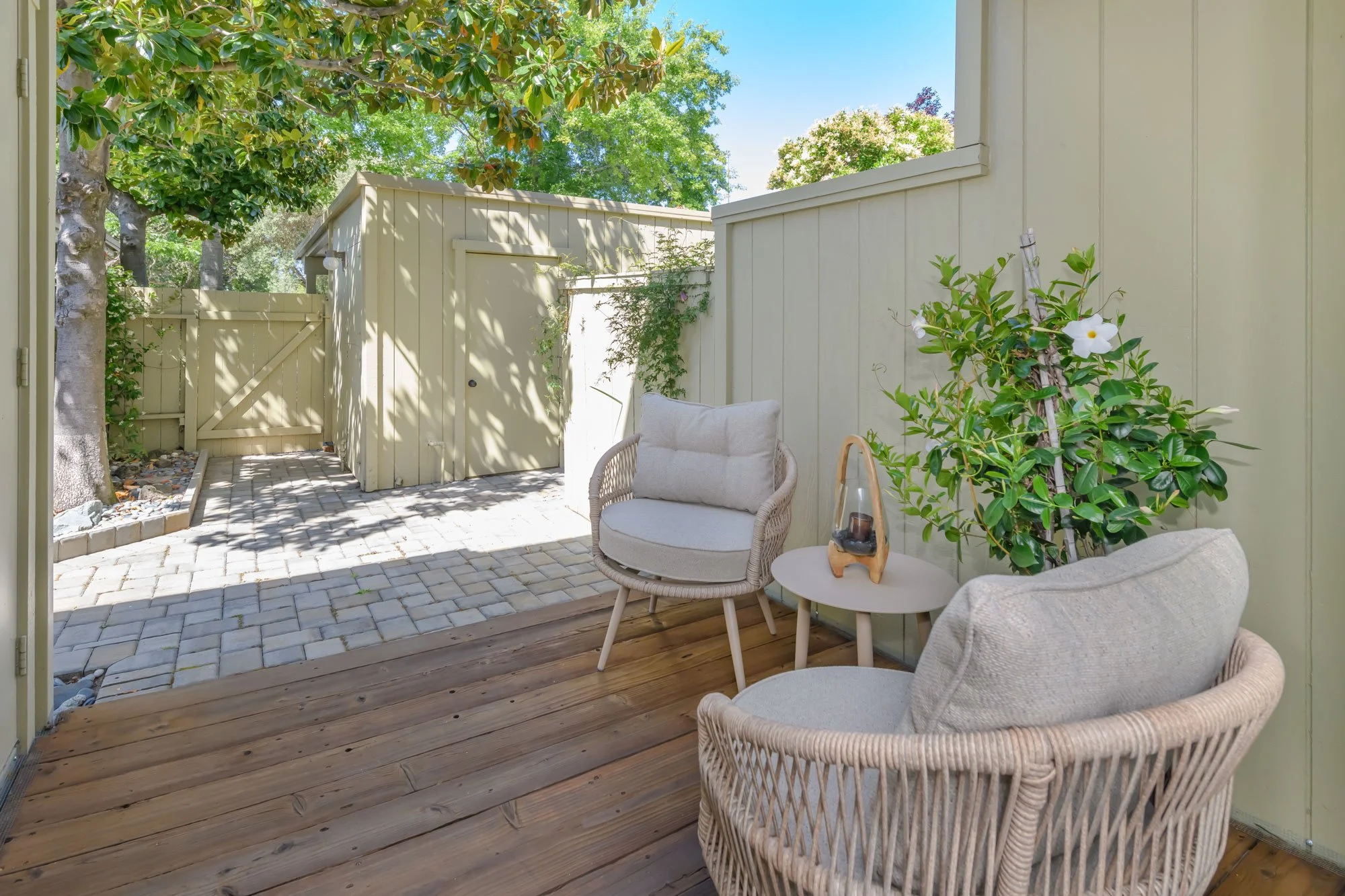 A cozy patio with wicker armchairs and a small table, surrounded by a beige wooden fence, with lush green trees overhead and blooming plants, under a clear blue sky.