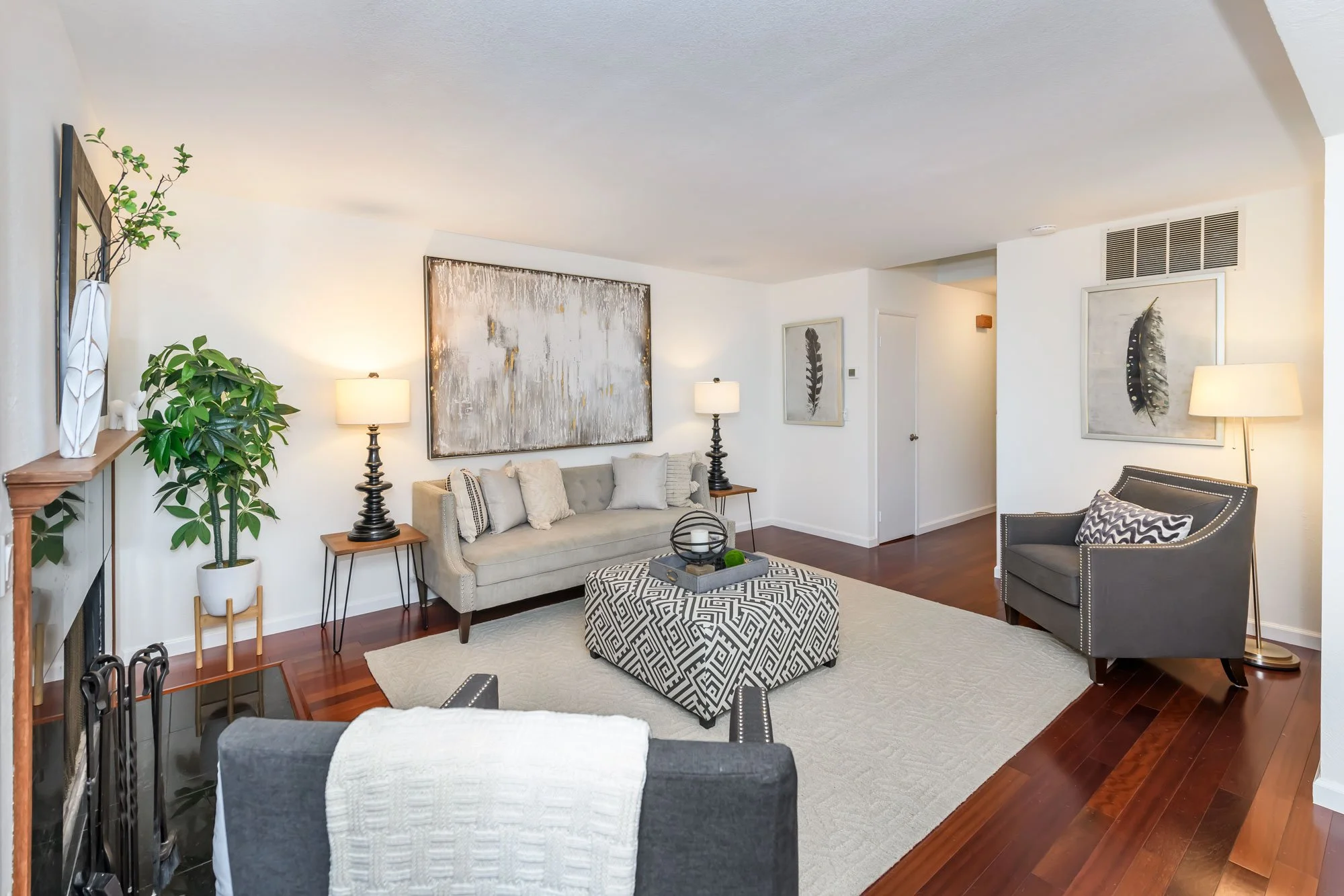Living room with a gray sofa, black side tables with lamps, a patterned ottoman, framed artwork of feathers on white walls, green plants, and hardwood floors.