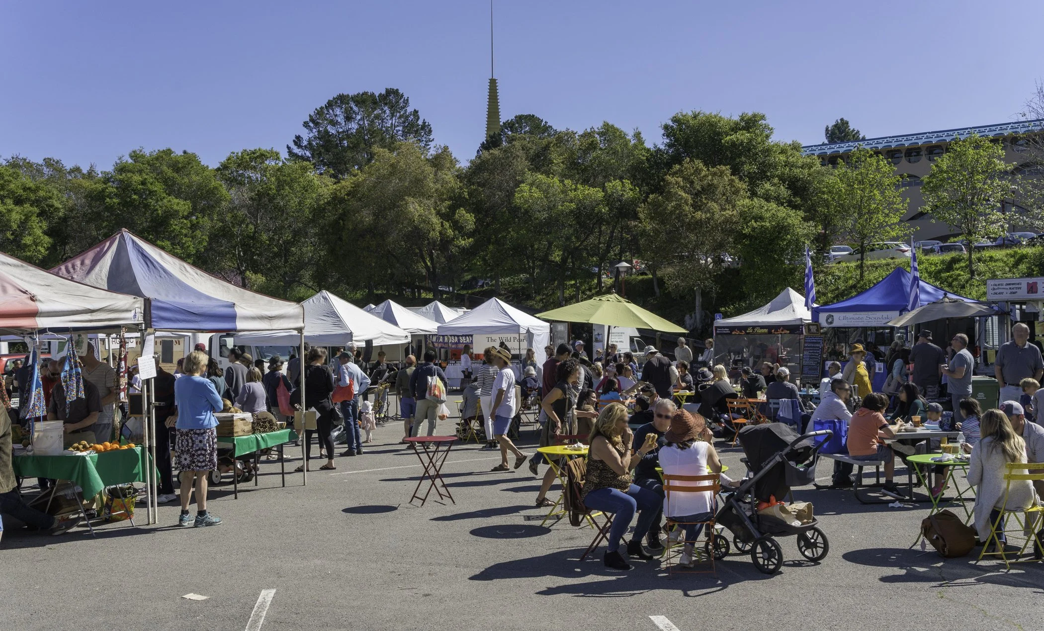 A busy outdoor market with white tents, vendors, and shoppers mingling. People are sitting at tables, walking, and browsing. Green trees and a building with a blue roof are in the background on a sunny day.