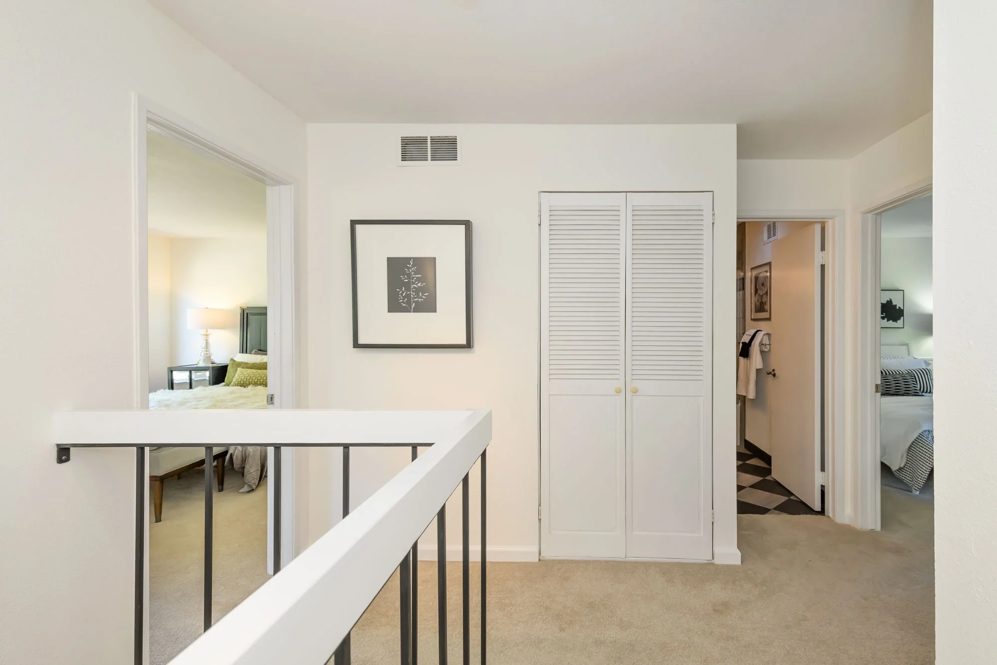 Interior view of a home featuring a hallway with a closet with louvered doors, framed artwork on the wall, and bedrooms visible through open doorways, with beige carpeting and white walls.