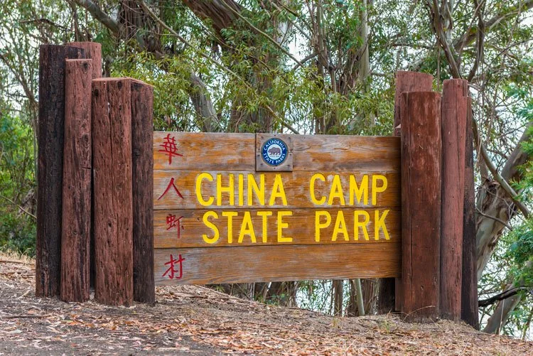 Wooden sign for China Camp State Park with yellow lettering on a natural setting with trees in the background.