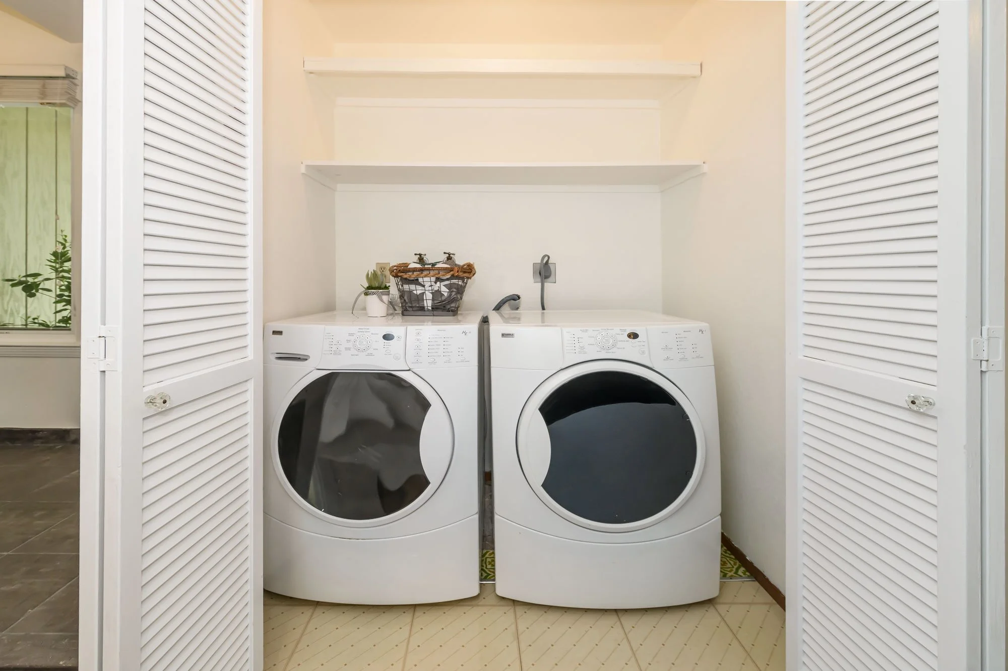 A laundry room with a white washing machine and dryer side by side, seen through open white louvered closet doors. There are three white shelves above the appliances, a small plant, and a laundry basket on top of the washing machine. The room has bei