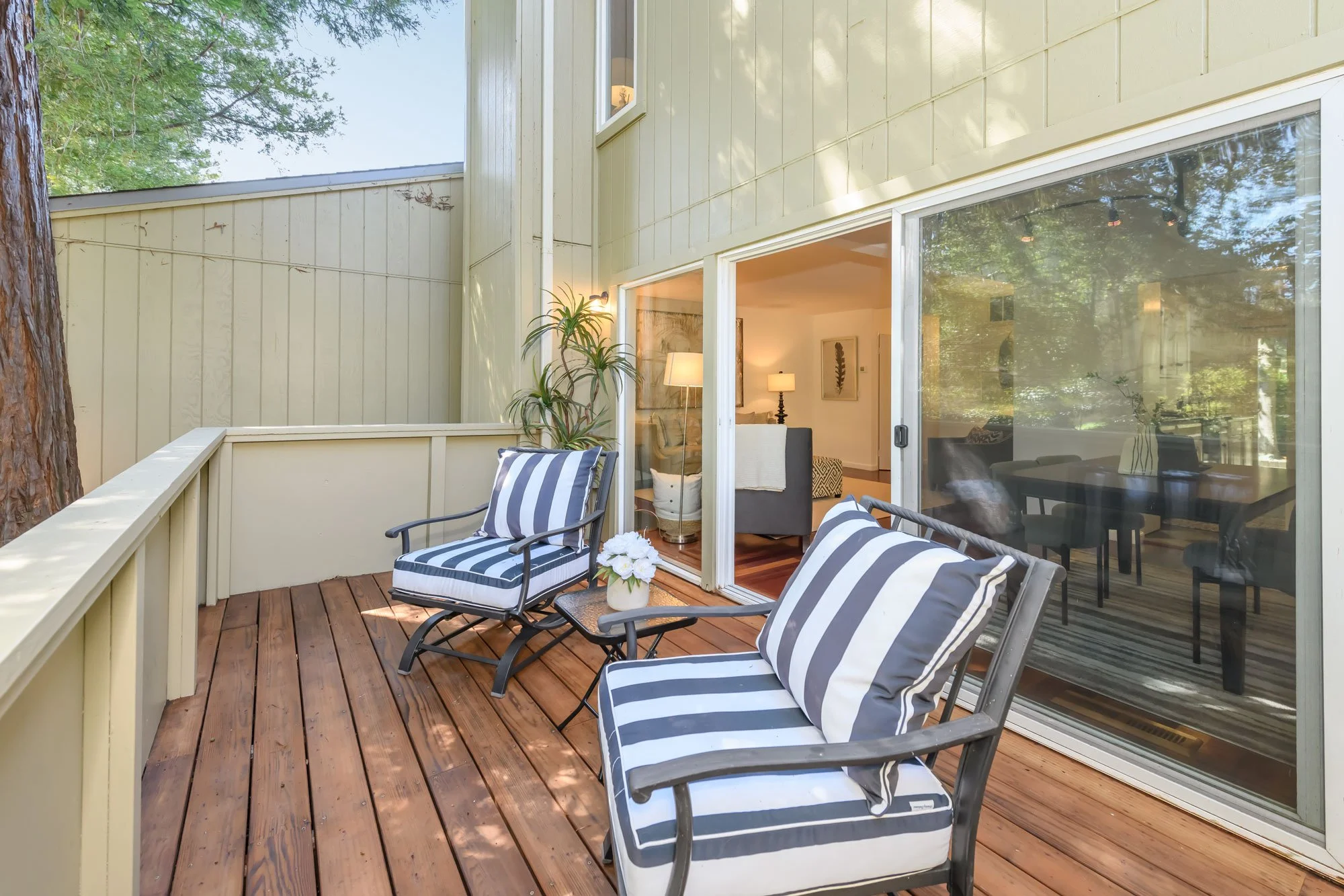 A cozy outdoor balcony with wood flooring, two armchairs with black frames and black and white striped cushions, a small table with a white flower arrangement, and an interior view of a living room with sofas, lamps, and artwork visible through glass