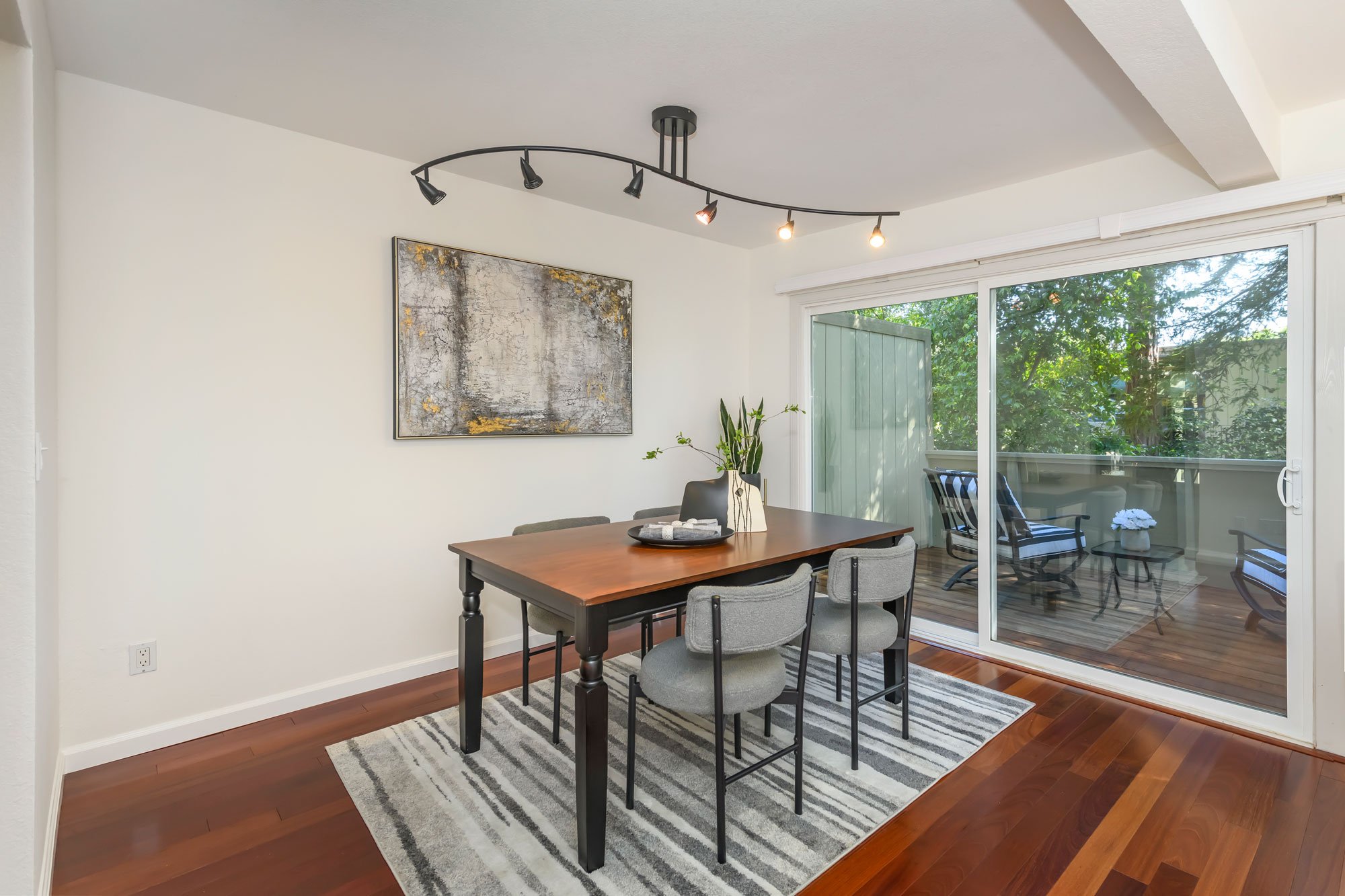 Dining area with a wooden table, four upholstered chairs, a striped rug, a modern picture on the wall, a sliding glass door leading to a balcony with outdoor seating and greenery outside, and a ceiling track lighting fixture.