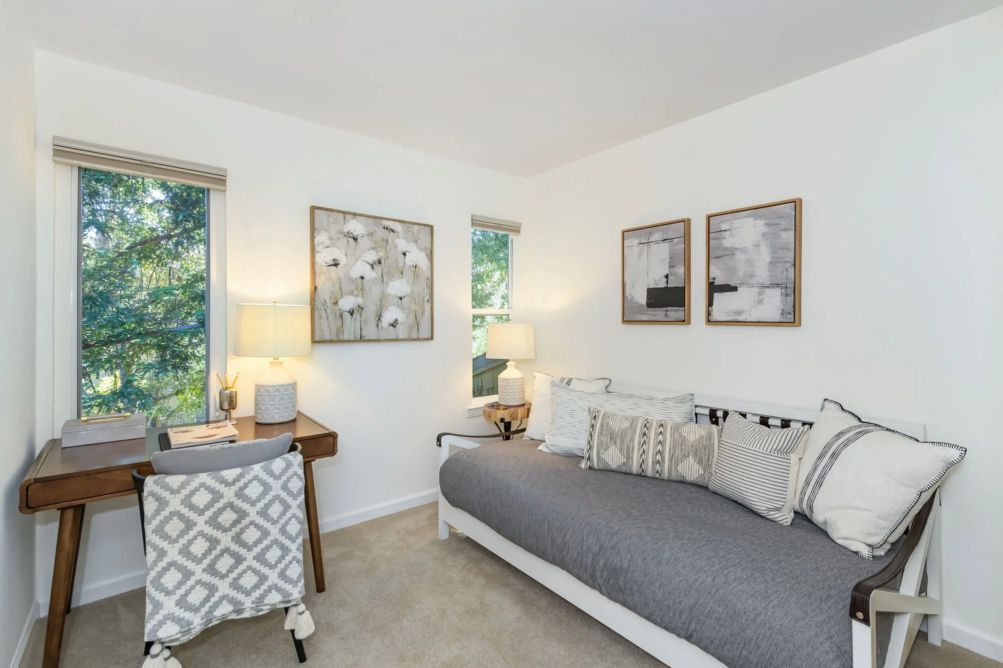 A cozy living room with a gray sofa topped with striped and patterned pillows. A side table with a lamp and decorative items is near the window, which shows green trees outside. The wall has three modern abstract paintings, and a wooden desk with a p
