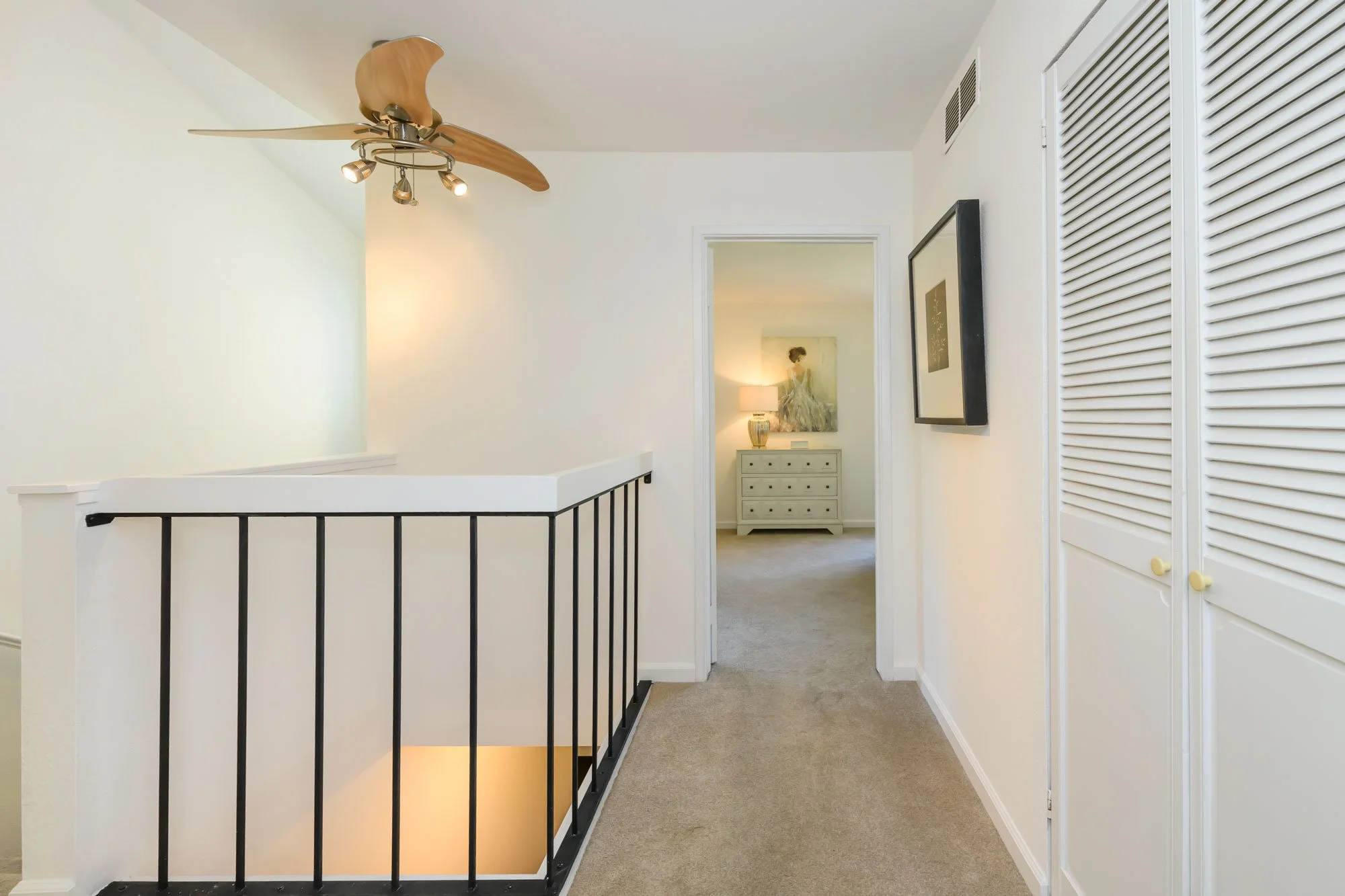 Interior view of a house hallway with beige carpet, white walls, a black metal railing, and a ceiling fan with wooden blades. Doorways lead to other rooms, one with a framed picture and a dresser illuminated by a lamp.