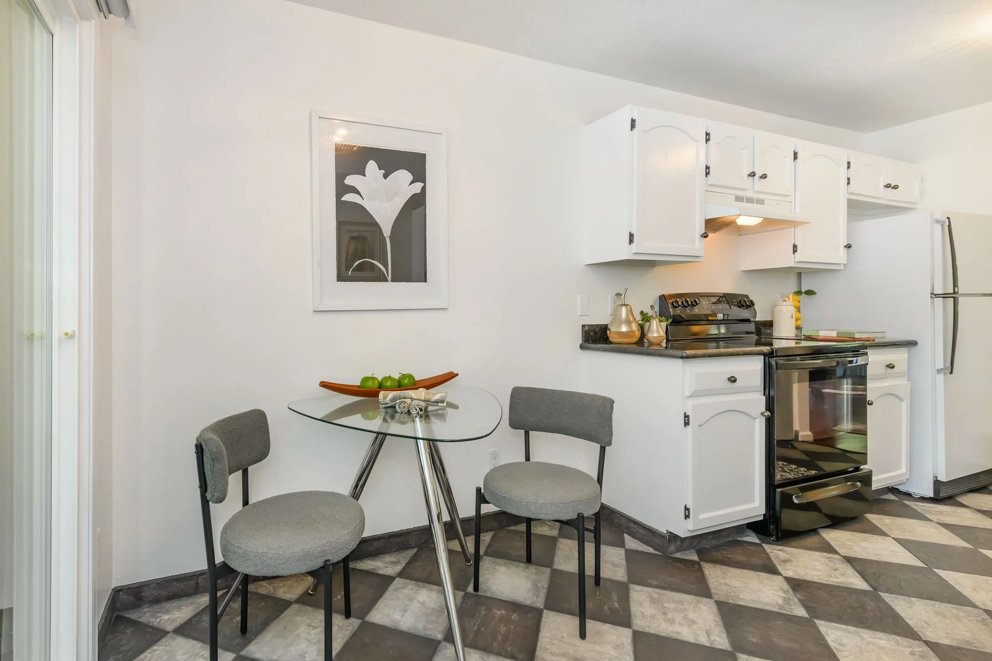 Kitchen with white cabinets, stove, refrigerator, and small dining table with three gray chairs, decorative items, and a framed flower picture on the wall.