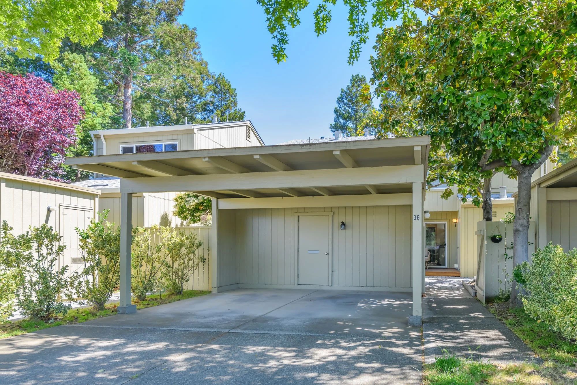 A beige residential building with a covered carport, surrounded by trees and greenery, under a clear blue sky.