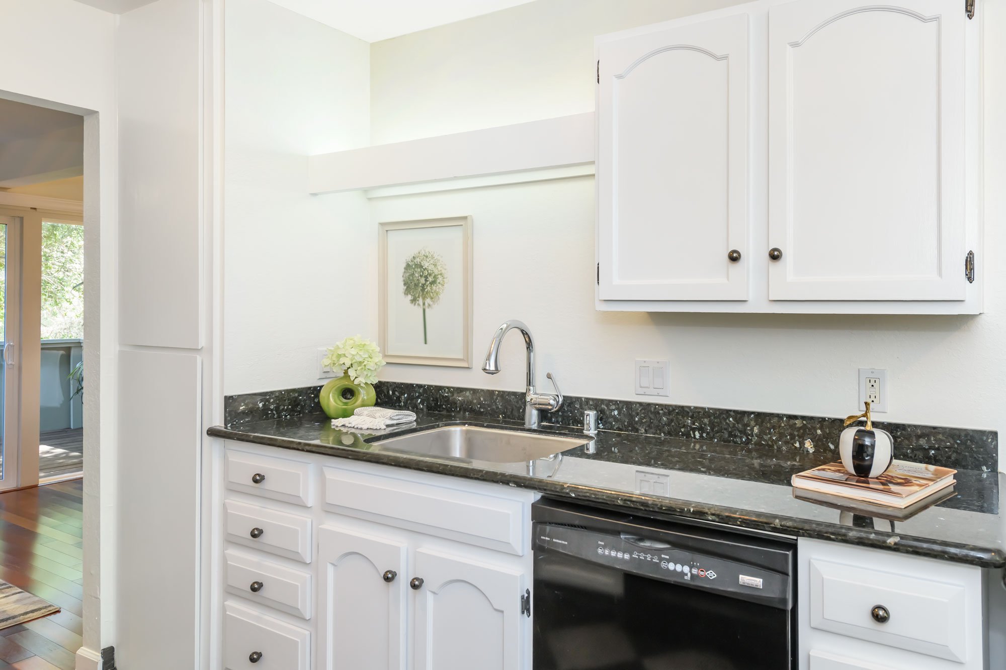 A white kitchen with white cabinets, a black speckled granite countertop, and a stainless steel sink with a chrome faucet. Decorative items include a green vase with white flowers, a framed botanical print, a small white and black speaker, and a stac