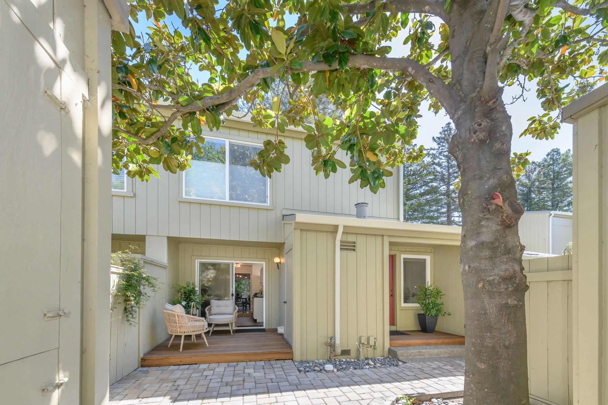View of a courtyard with a large tree, a sunny sky, and a two-story beige house with windows. The outdoor area has a patio with chairs and plants.