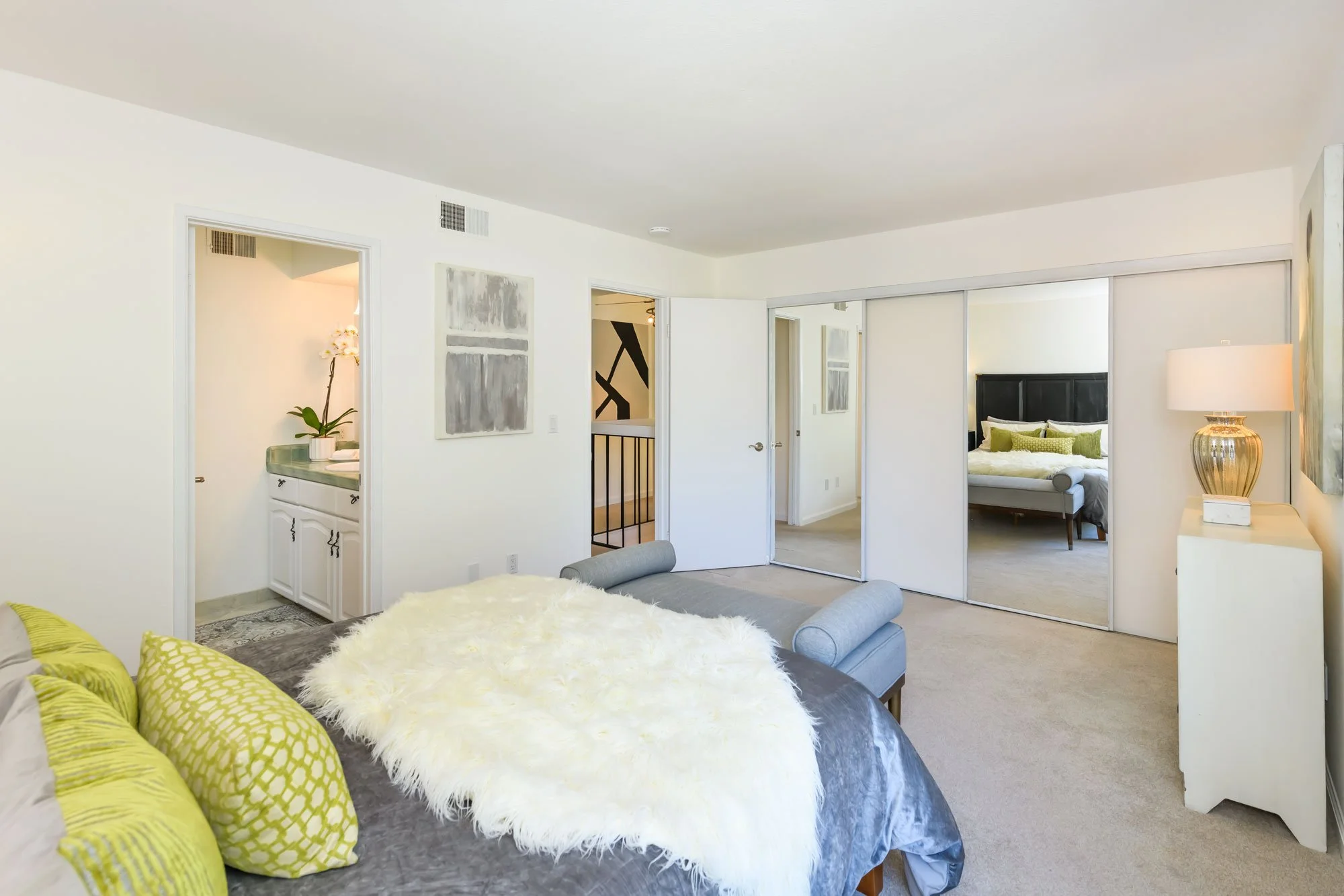 A bedroom with a bed, green pillows, a furry throw, and a mirror closet door reflecting the room. An adjacent room has a nightstand with a lamp, artwork on the walls, and a small bathroom or vanity.