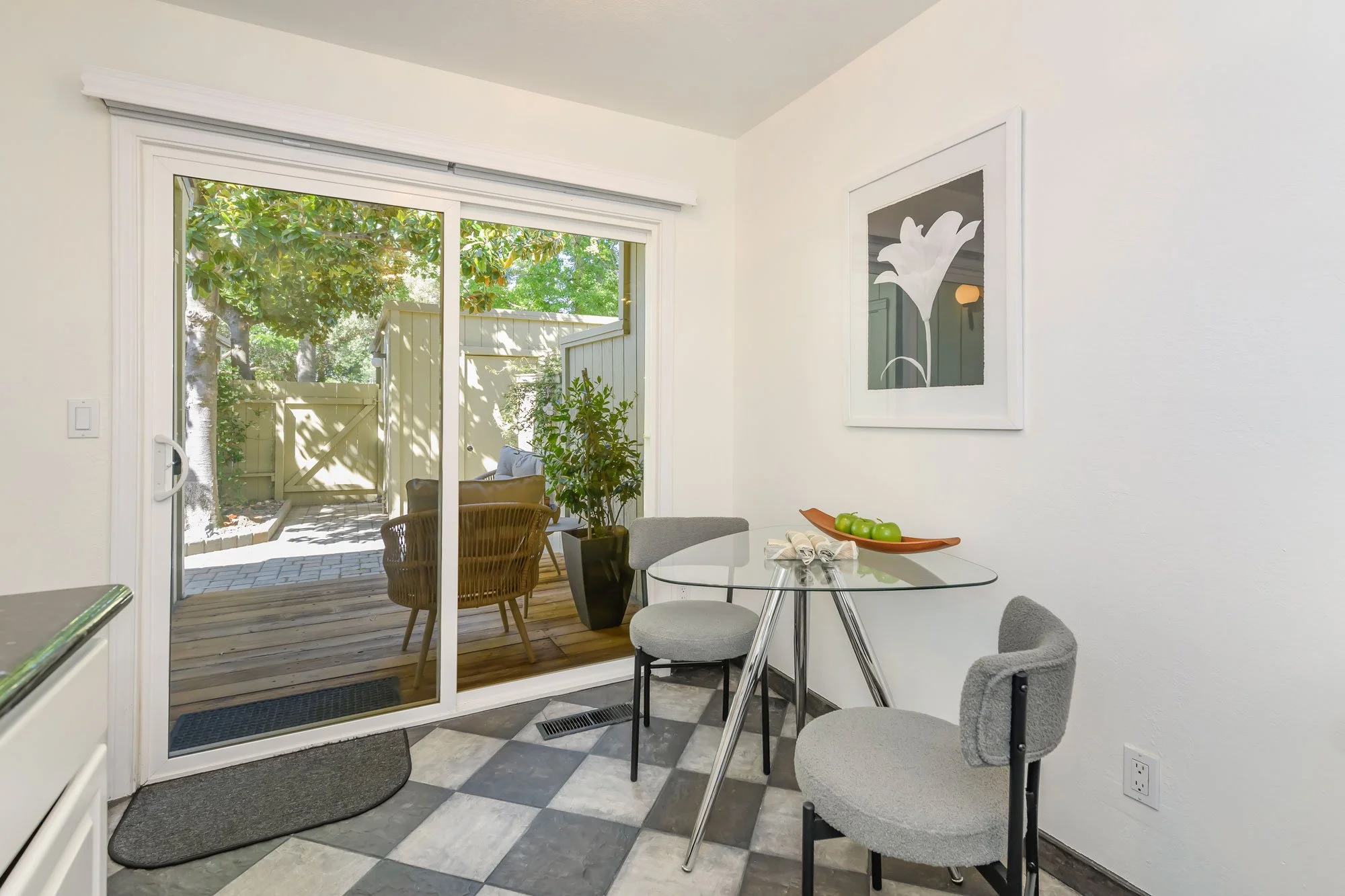 Interior view of a dining area with a glass round table, chairs, and a sliding glass door leading to an outdoor patio with trees and greenery.