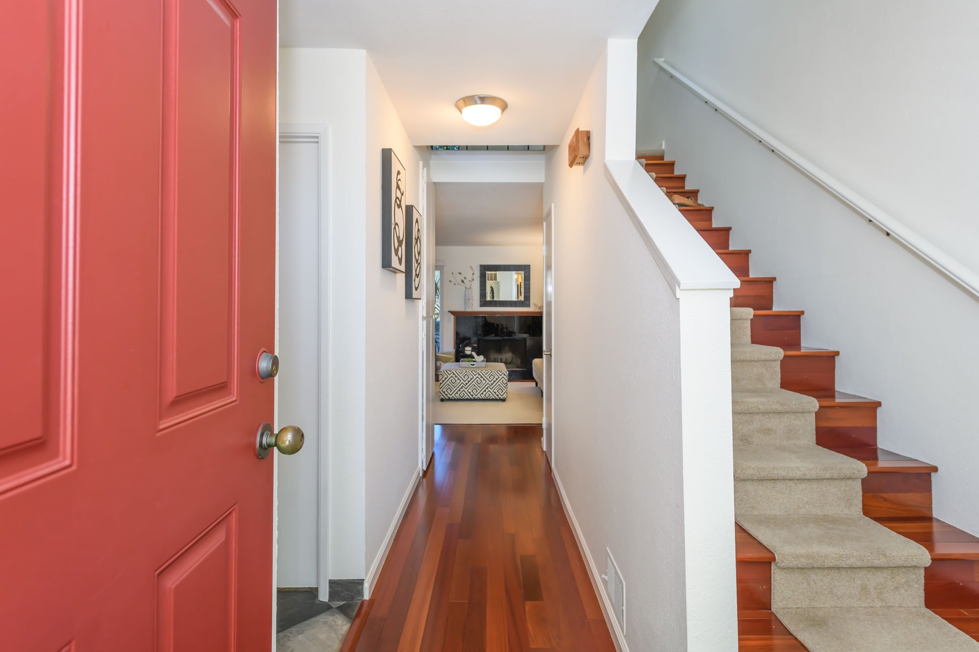 Interior view of a house entryway with a red front door, hardwood flooring, white walls, and a staircase with carpeting leading upstairs. Decor includes artwork on the walls and a mirror reflecting a living room area.