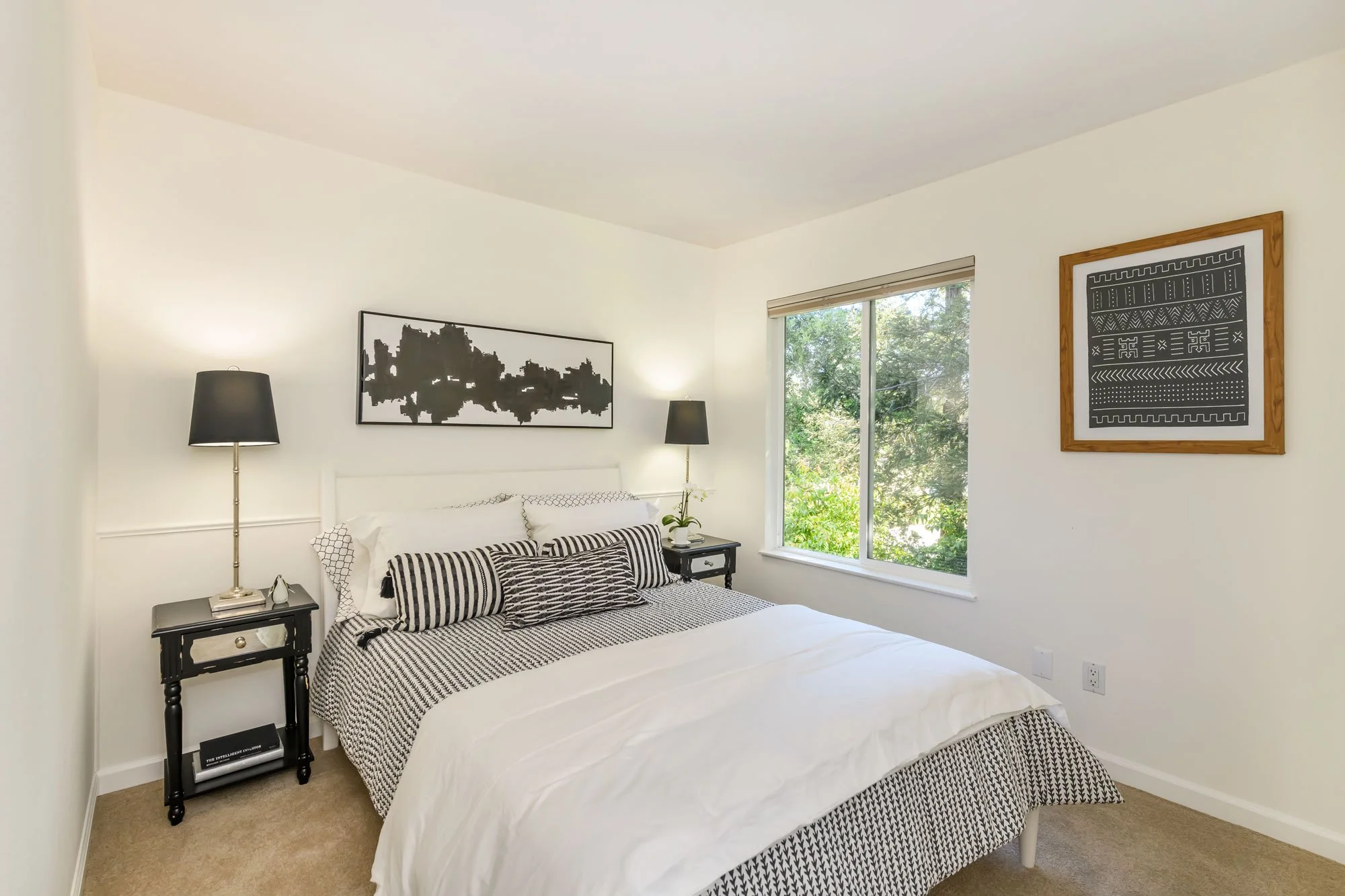 A bedroom with a white bed and black-and-white striped pillows, black nightstand with books and a lamp, wall art with black and white patterns, a window with a view of green trees, and beige carpet.