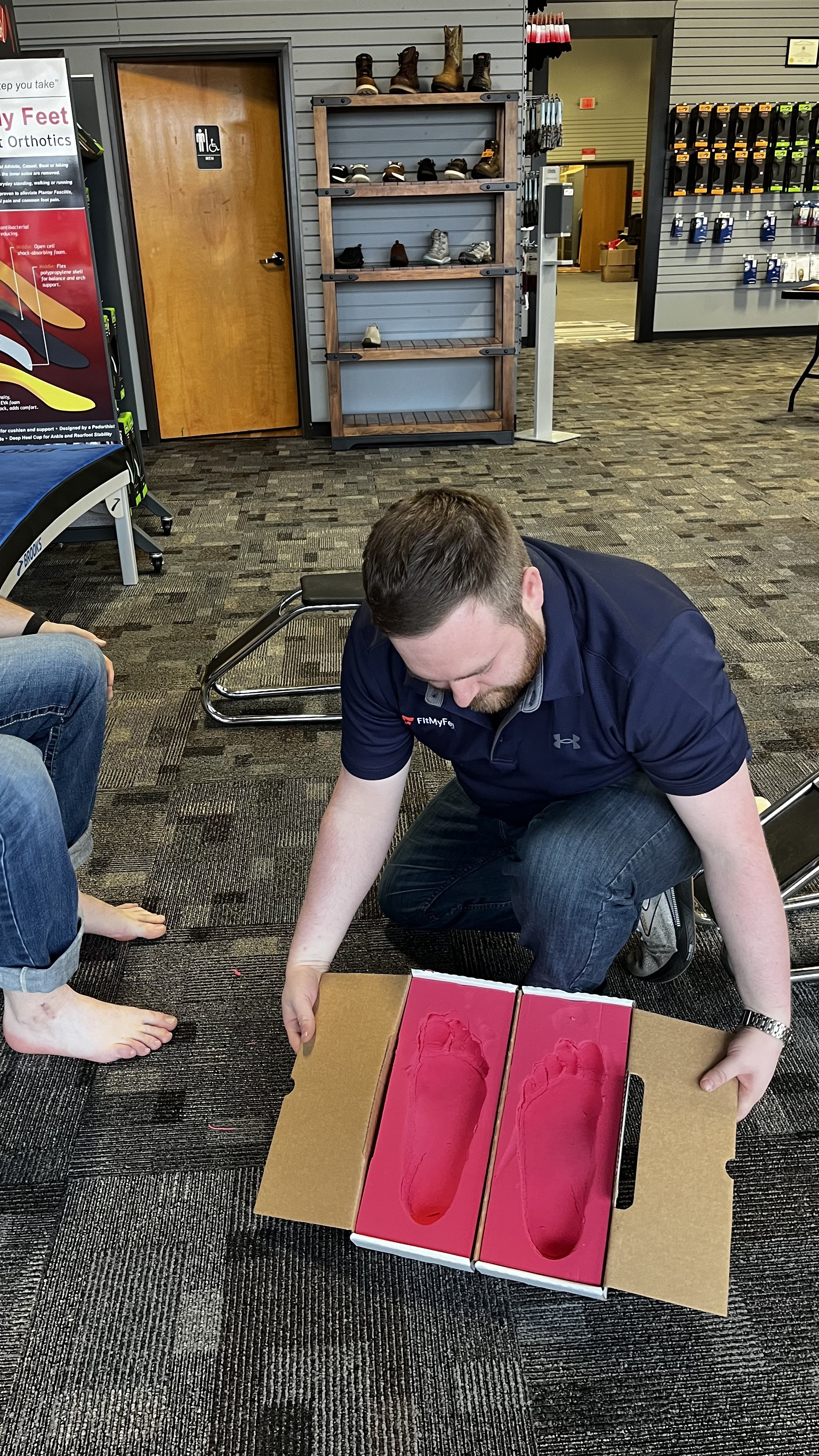 A man kneeling on the floor holding a custom orthotic box with red foam inside, next to another person who is barefoot sitting in a chair.