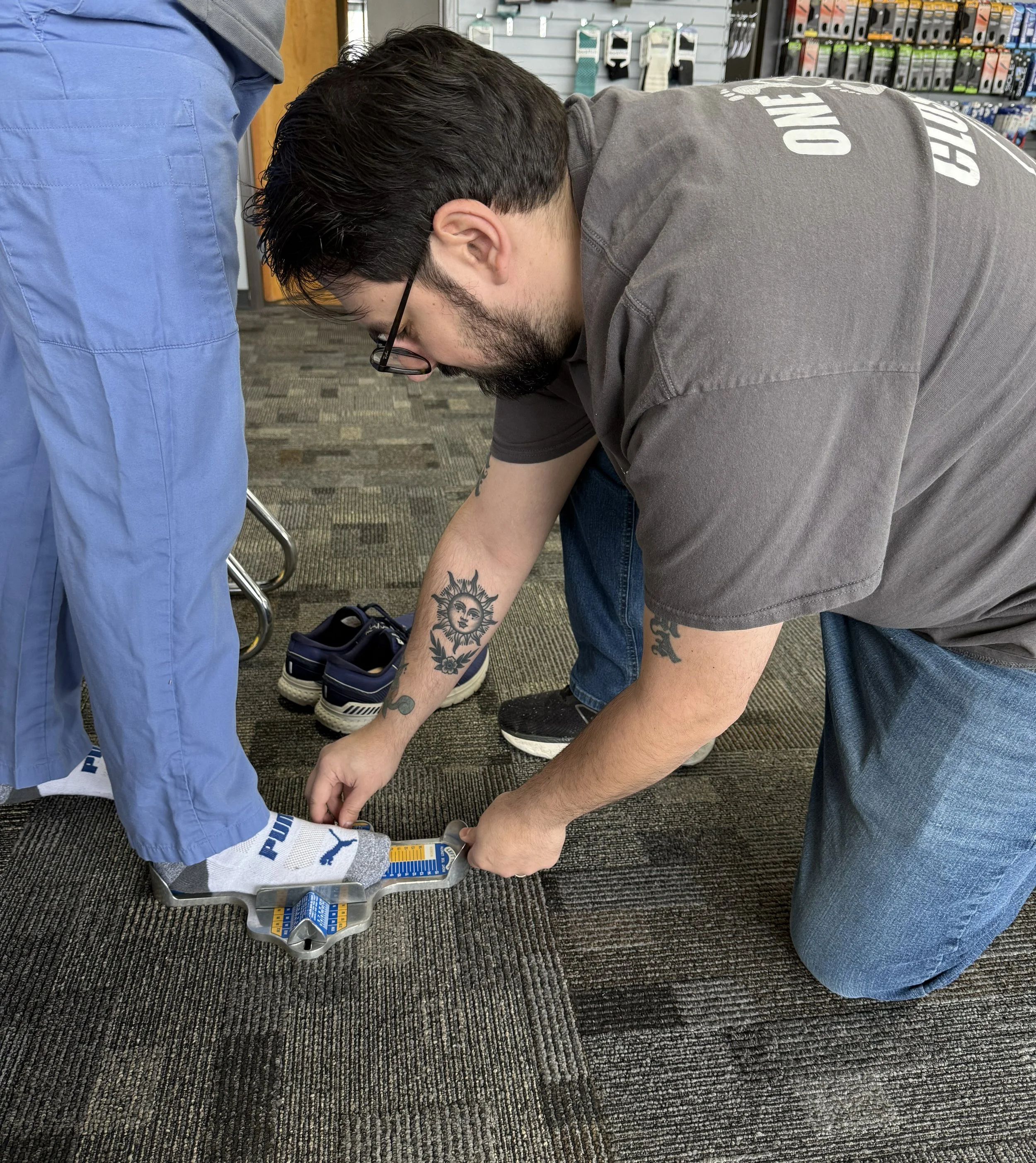A man kneeled over helping another custom by accurately measuring his foot length.