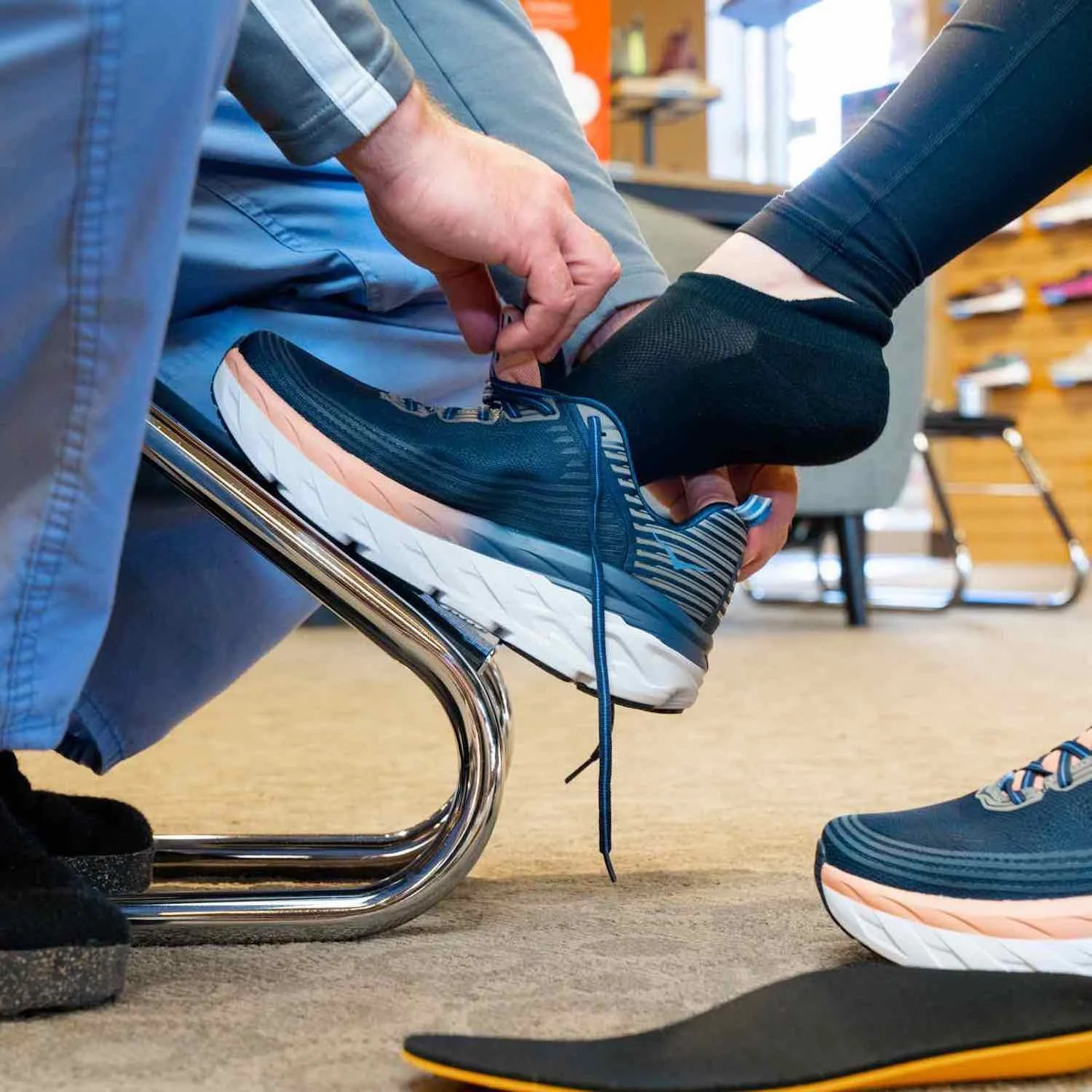 An employee helping a customer put their foot into a pair of blue colored shoes for accurate fit while there is a pair of over-the-counter insoles on the floor for extra support and cushioning in the new pair of shoes.