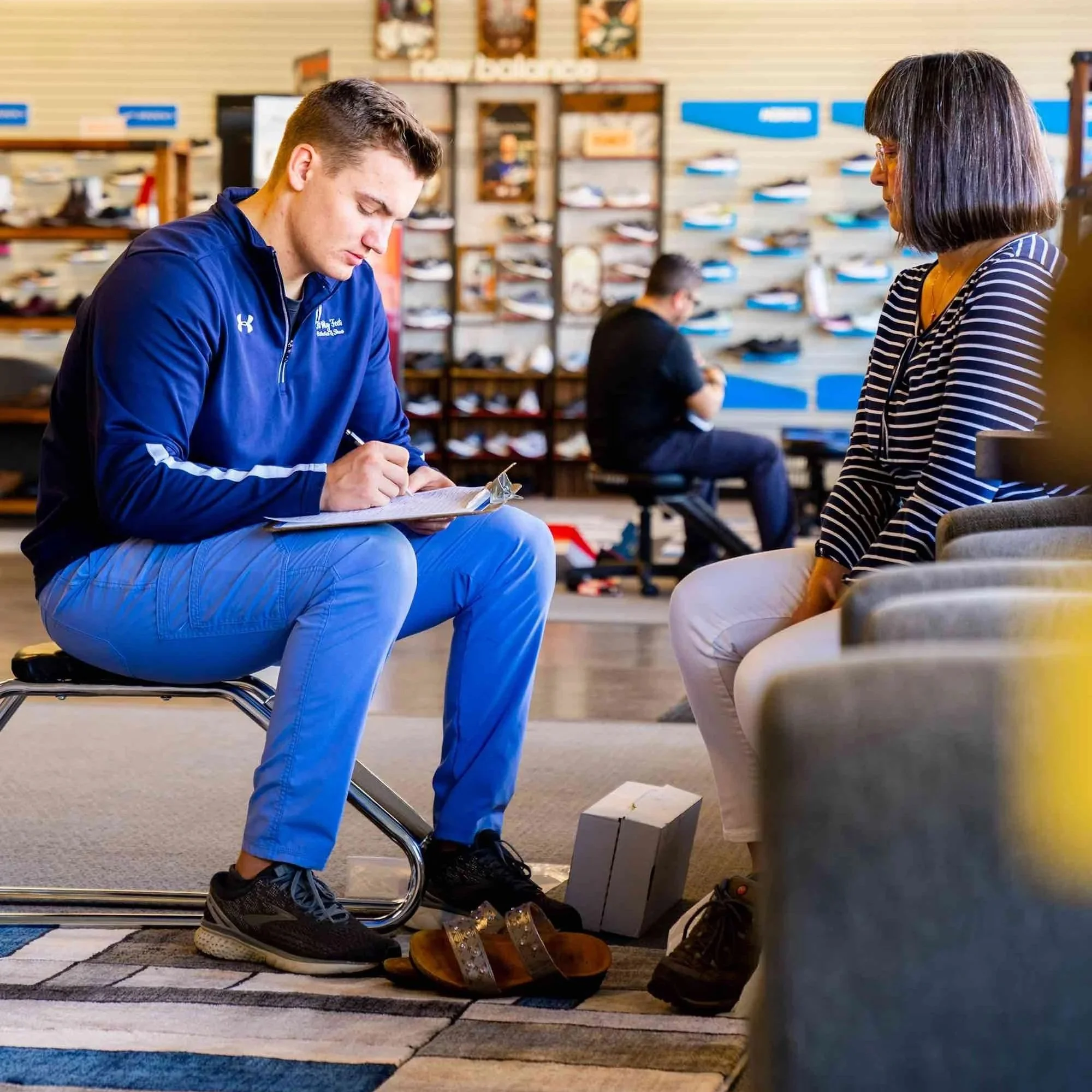 A young man in blue athletic clothing sitting on a chair, writing in a notebook, with a woman seated across from him in a shoe store or sports store.