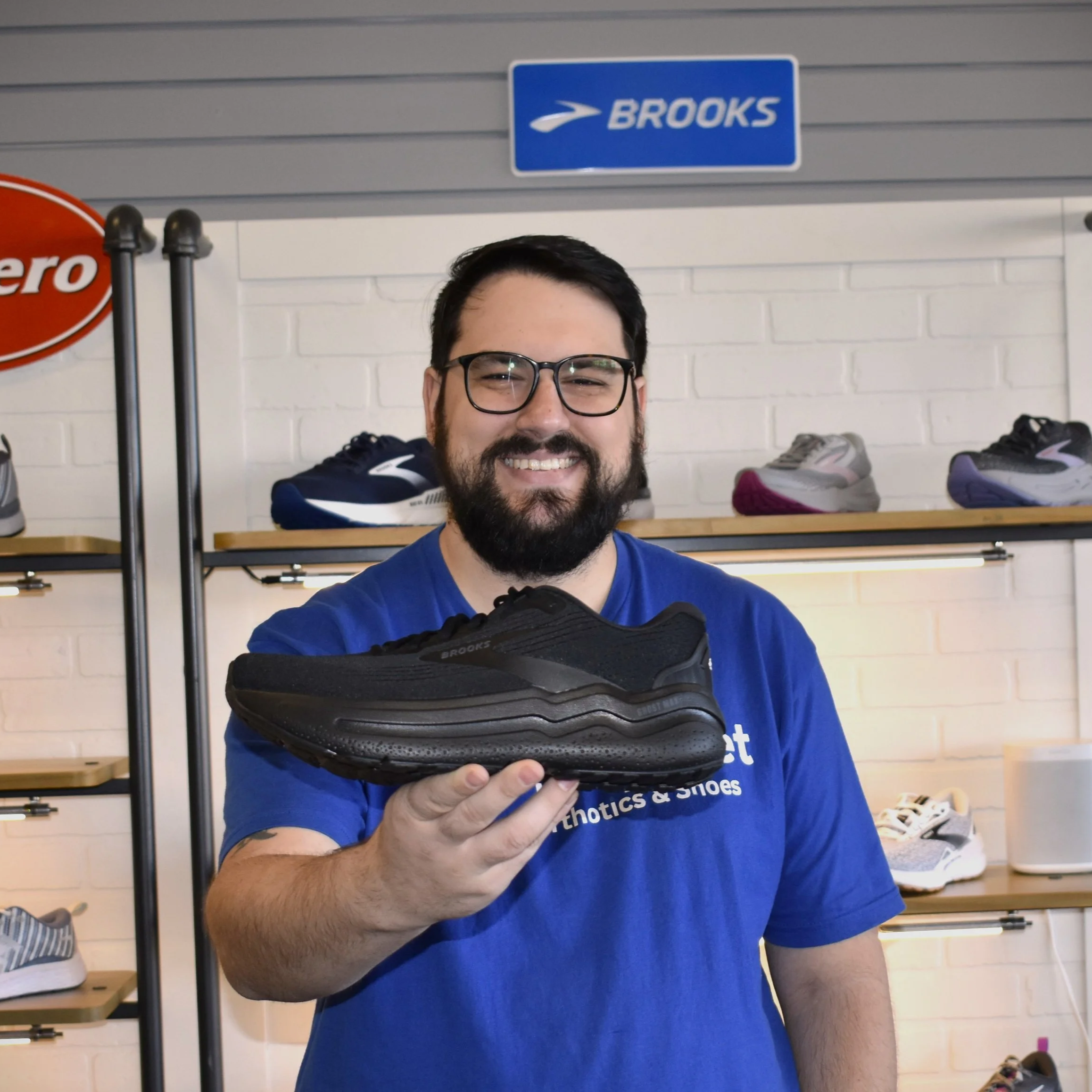 A man with glasses and a beard smiling and holding a black athletic shoe inside a sneaker store.