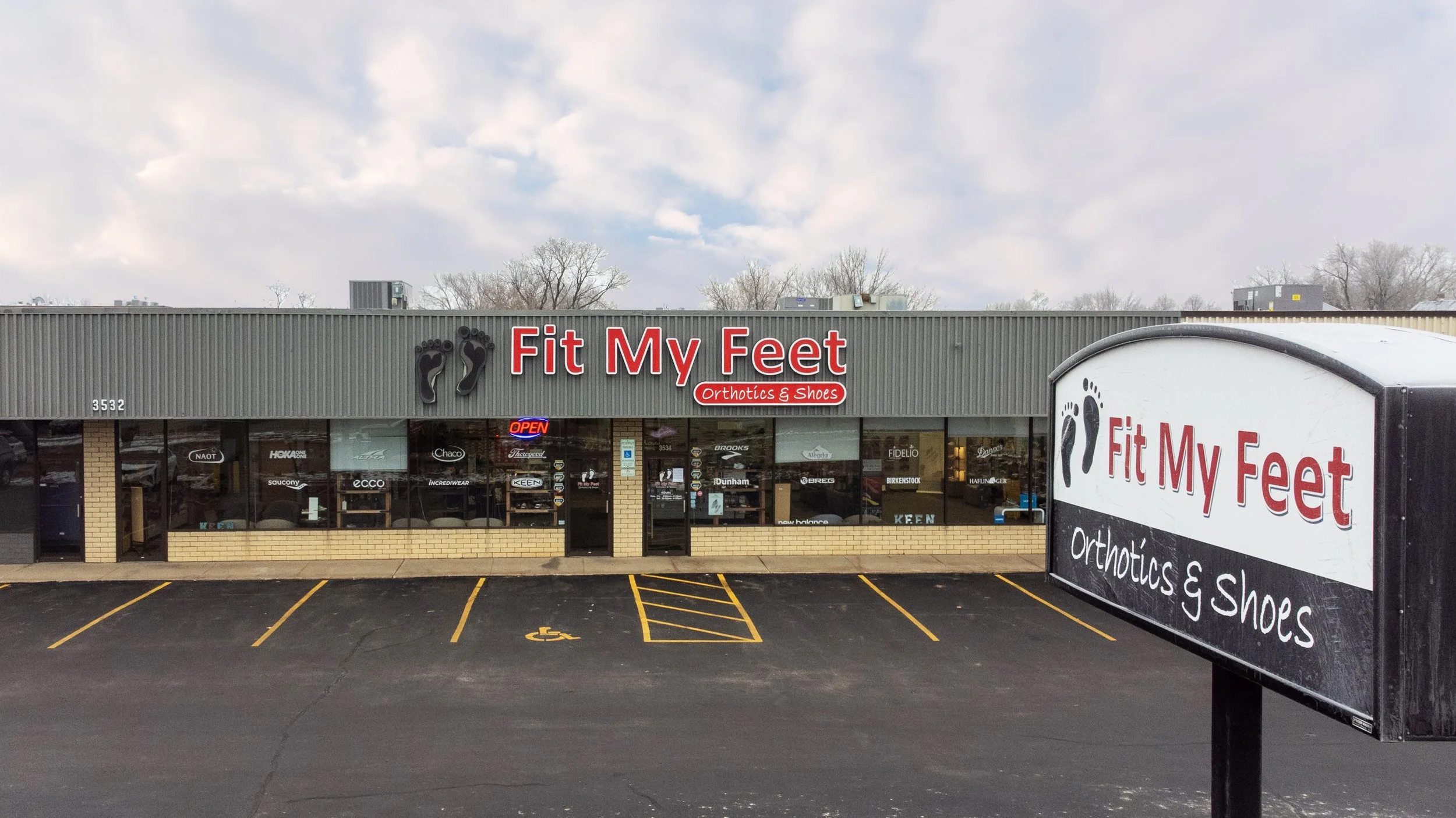 Exterior of a store named Fit My Feet Orthotics & Shoes with a parking lot in front, an open sign, and a large sign in the foreground displaying the store name and services.