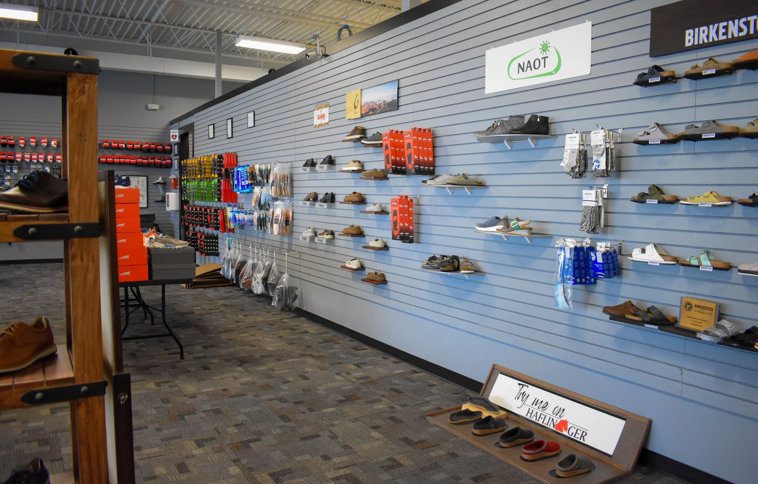 Shoes and accessories displayed on grey slatwall panels in a retail store, with promotional signs and a carpeted floor.