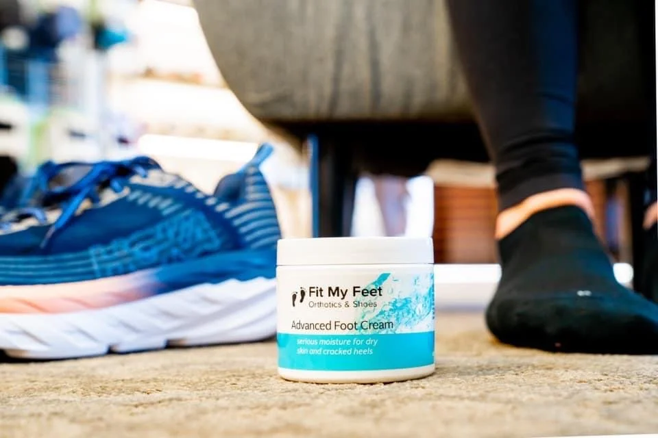 Close-up of a container of 'Fit My Feet' advanced foot cream on a carpeted surface, with a blue running shoe on the left and a person's foot on the right wearing a black sock.