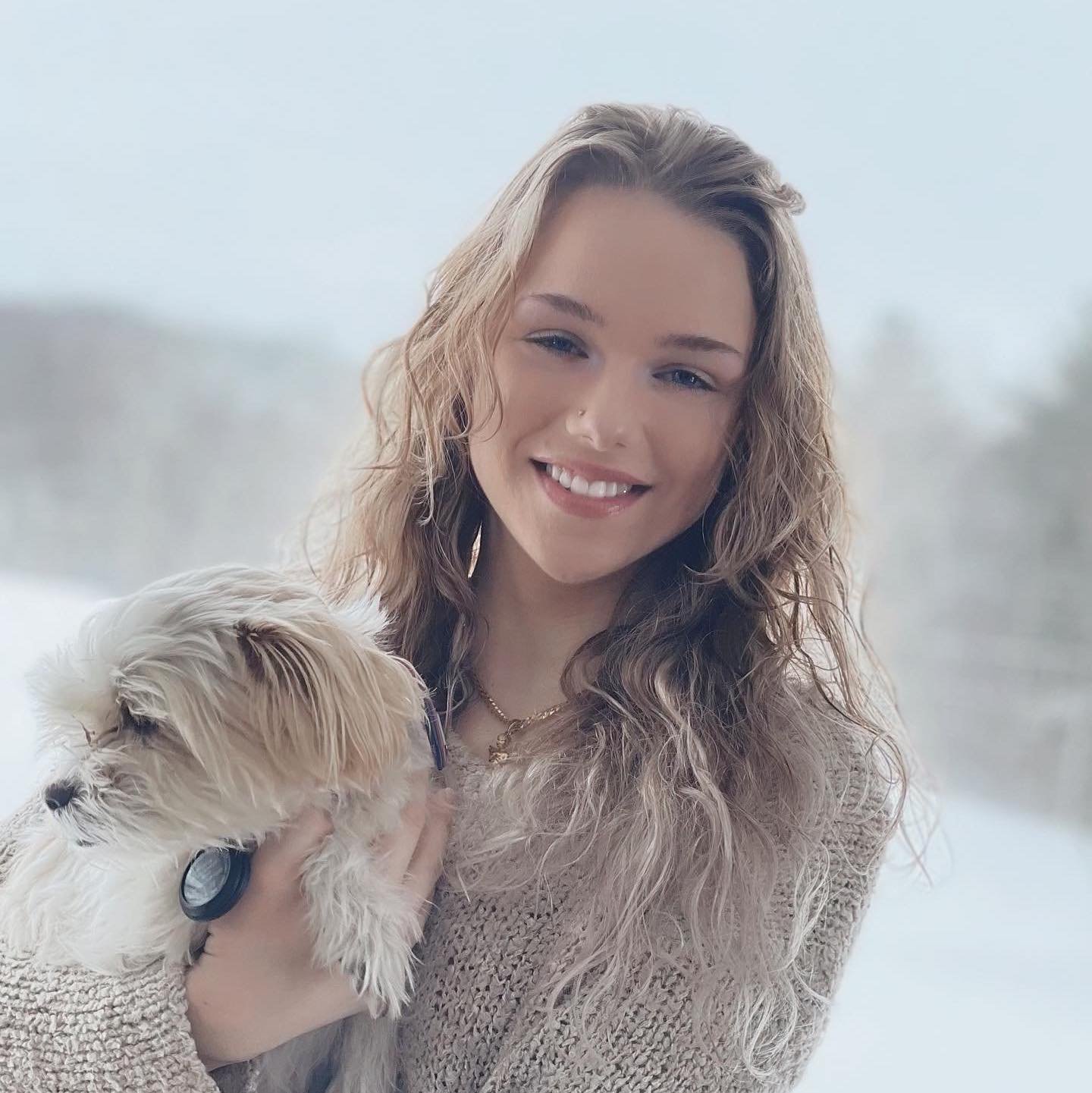 A young woman with wavy blonde hair smiling and holding a small fluffy white dog.