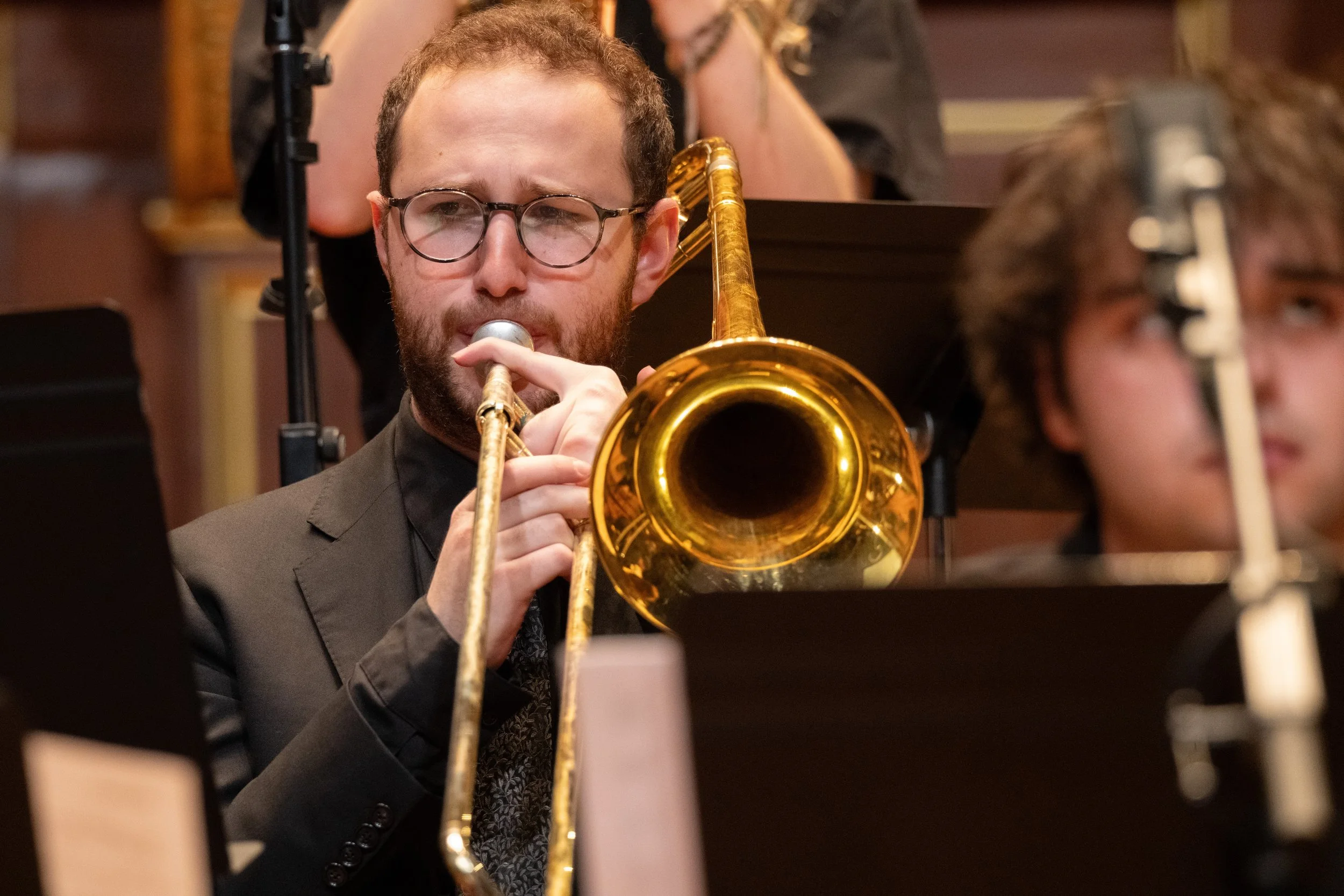 A young man with glasses and a beard playing a gold trumpet in an orchestra, with sheet music and other musicians nearby.