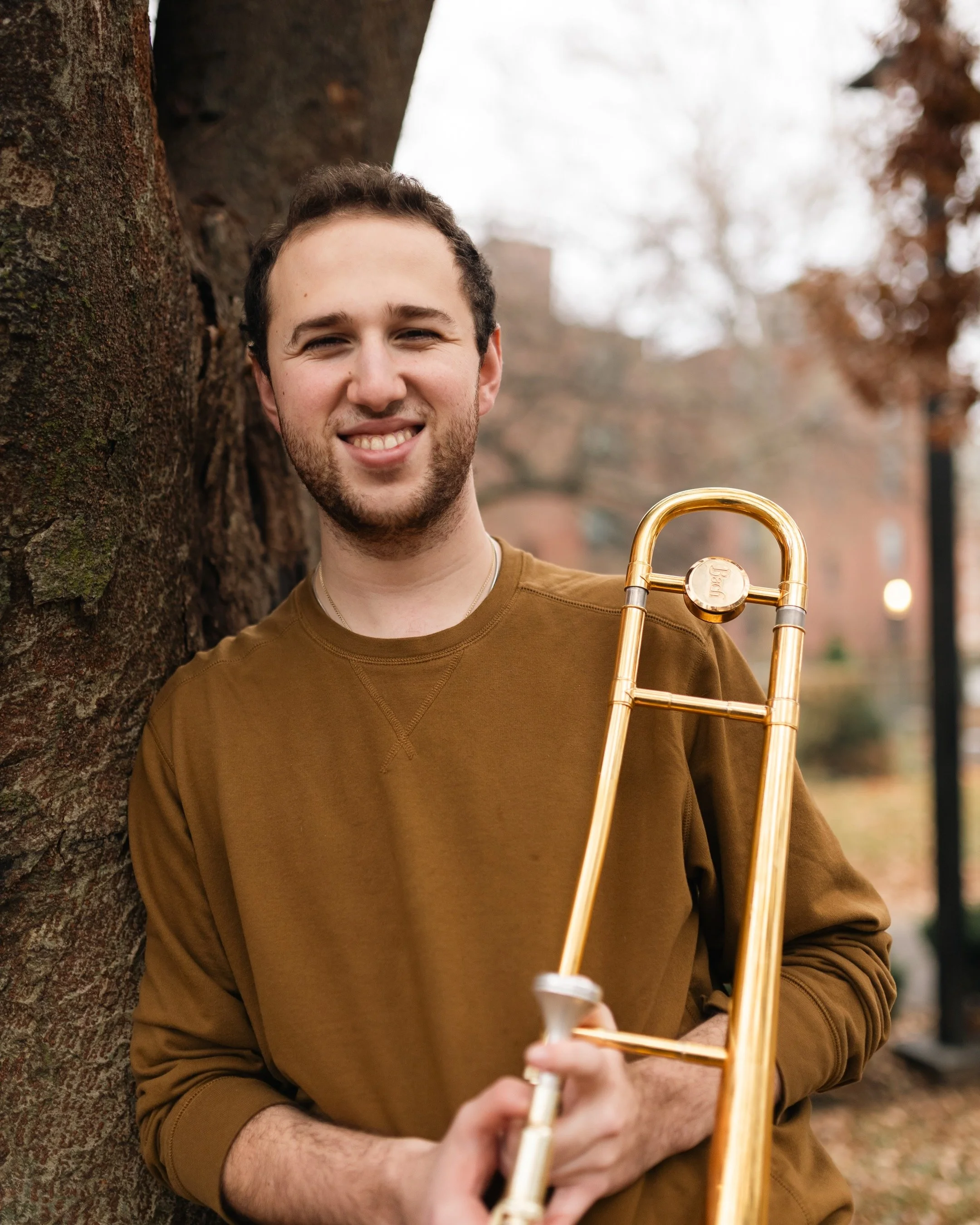 A young man with short brown hair and a beard, smiling, leaning against a tree in an outdoor setting with autumn leaves in the background, holding a trombone.
