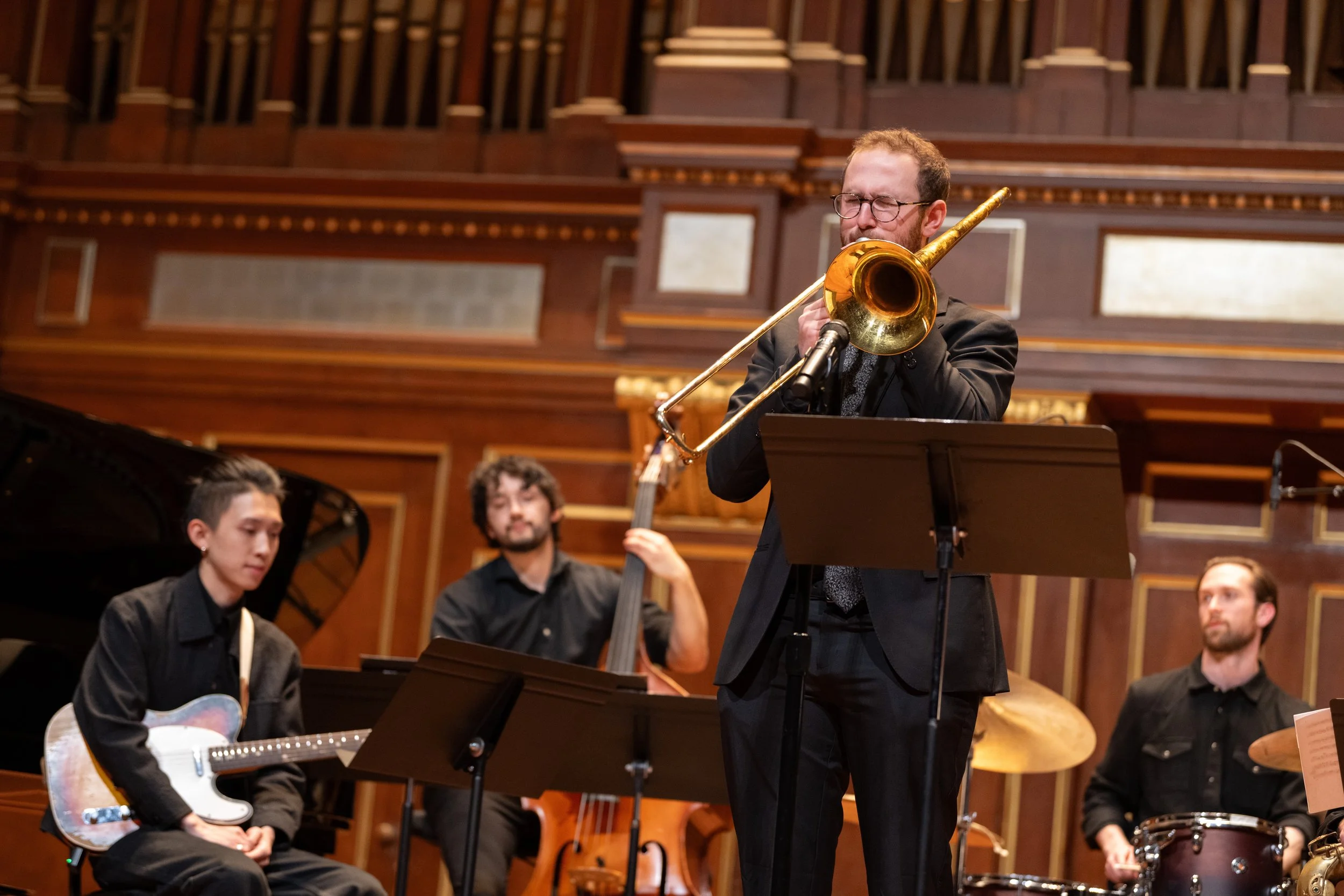 A man with glasses playing a trombone in an orchestra, with a group of musicians including a guitarist and other instrumentalists in a wood-paneled concert hall.