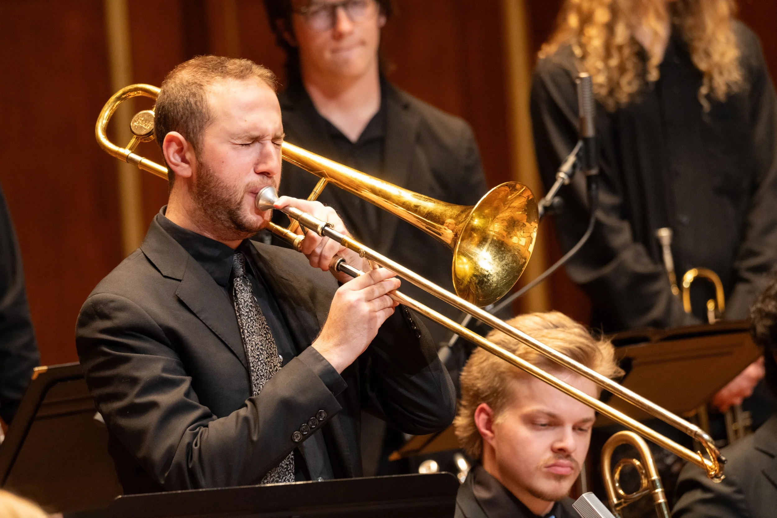 Musician playing the trombone during a concert with other orchestra members in the background.