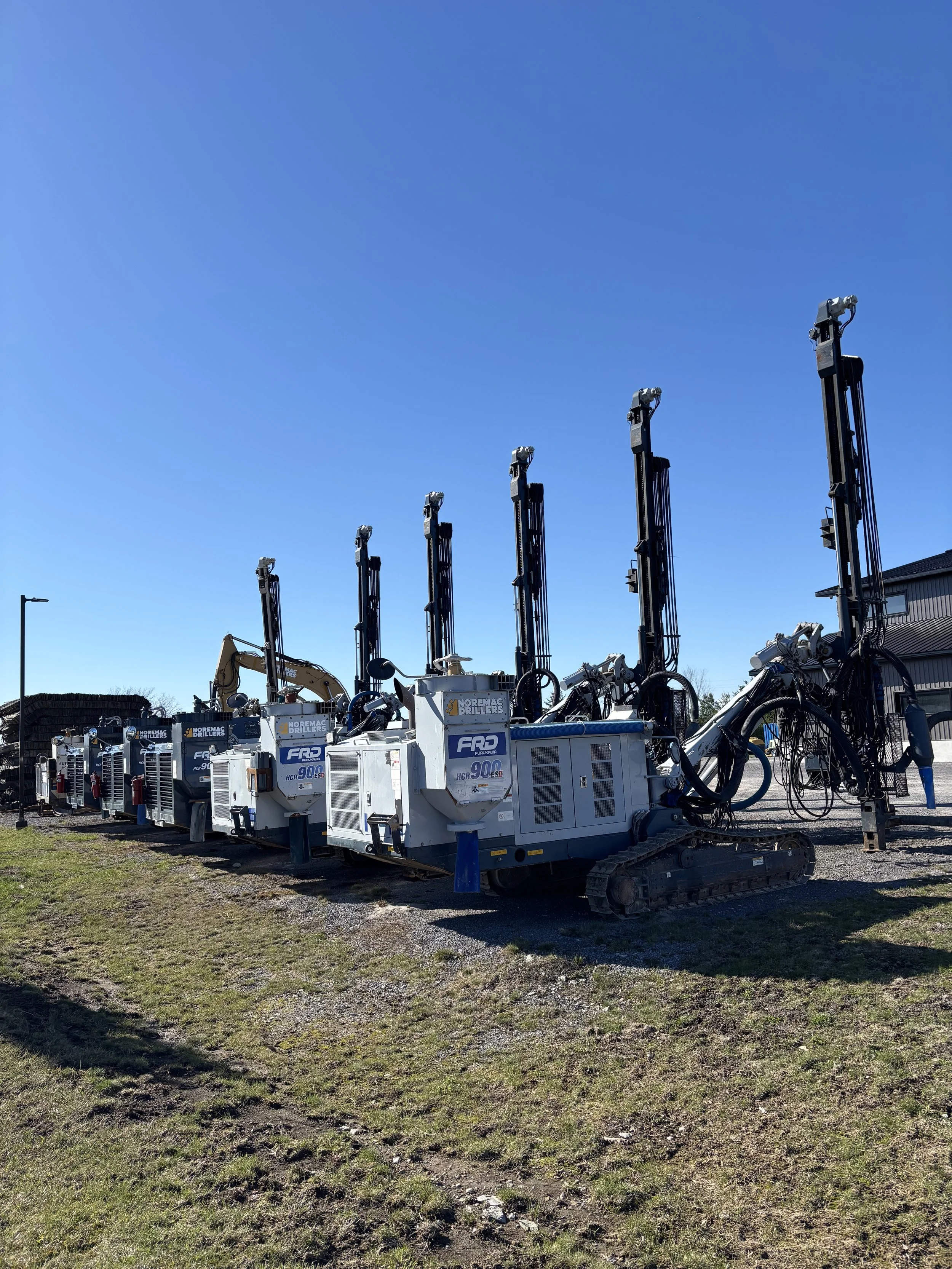 Row of large, industrial digging machines with long arms and tracks, parked outdoors on a grassy and gravel area under a clear blue sky.