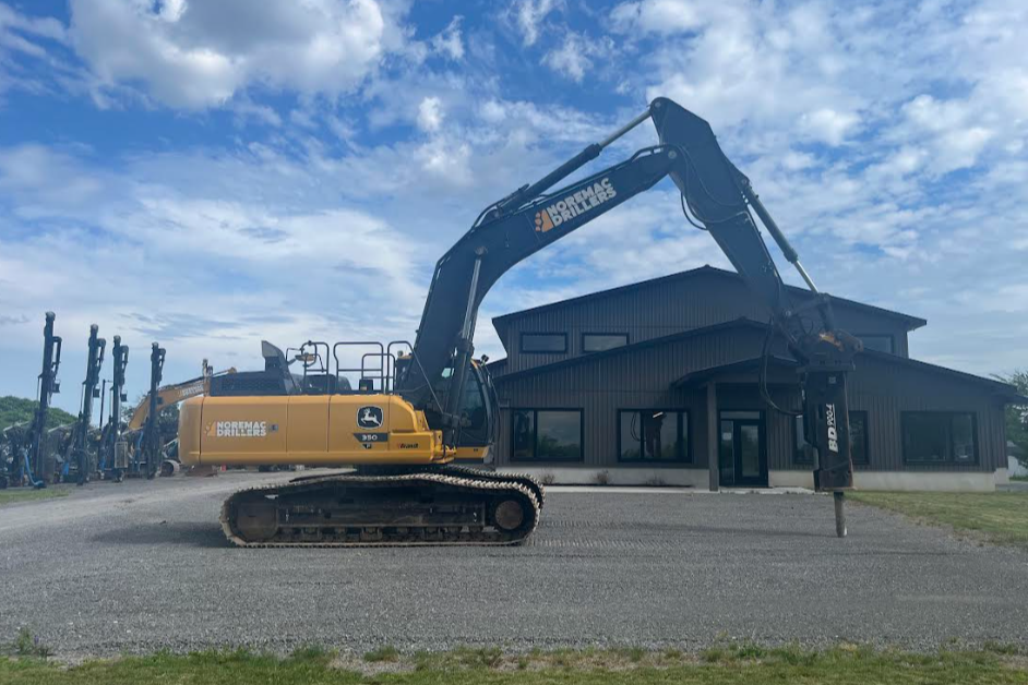 A large yellow excavator with black tracks parked in front of a dark gray industrial building on a gravel lot under a partly cloudy sky.