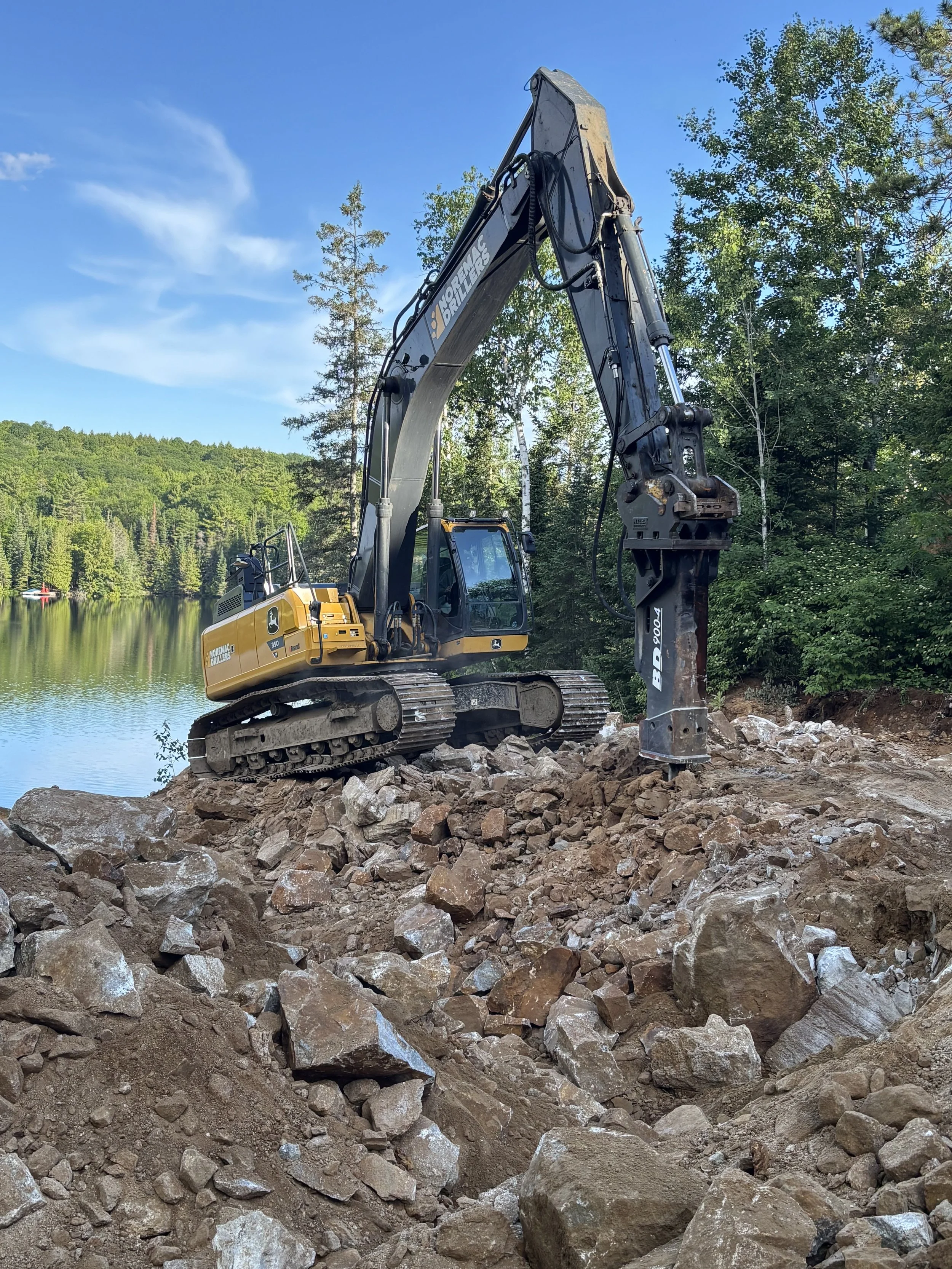 A large excavator working on rocky terrain near a body of water surrounded by trees, under a blue sky with some clouds.